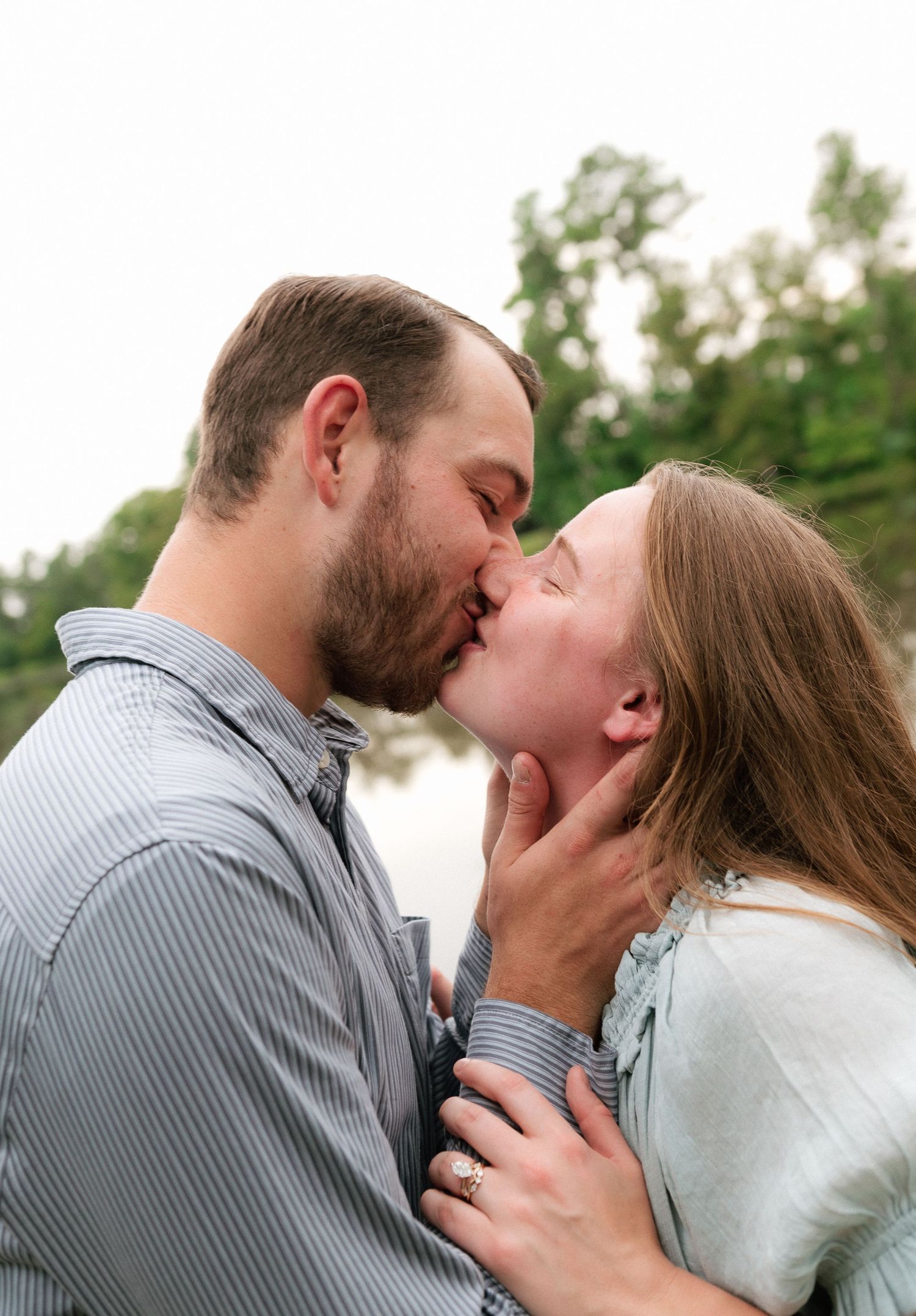 A couple shares a romantic kiss outdoors near a lake, surrounded by lush green trees in soft natural light.