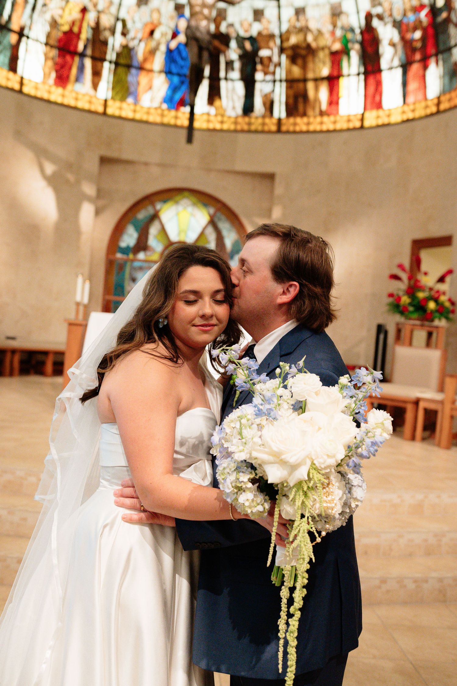 Bride and groom share a tender kiss in a church with stunning stained glass windows, bride holding white and blue bouquet.