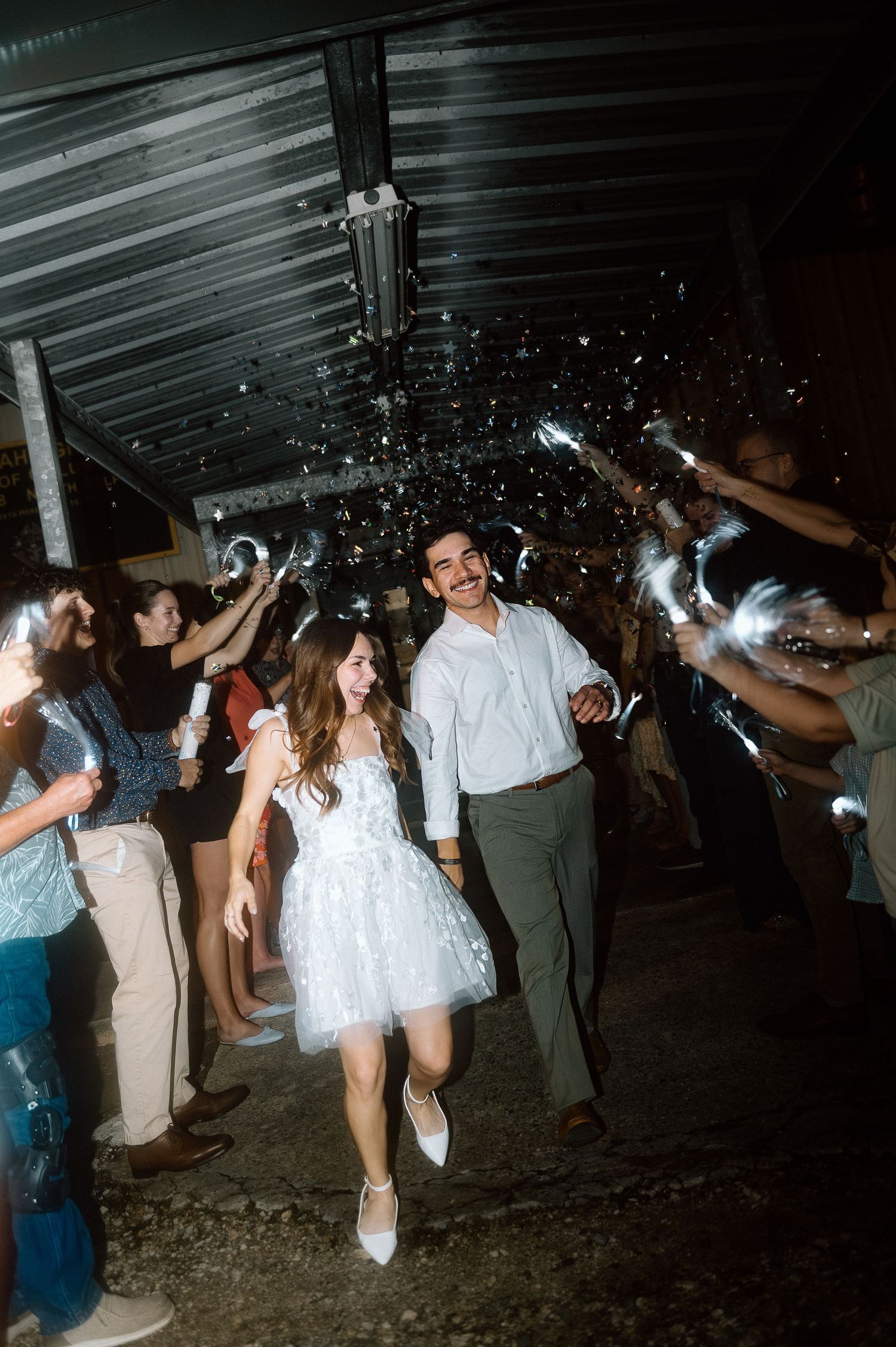 Wedding guests dance and celebrate with sparklers during a lively nighttime reception under a covered venue.