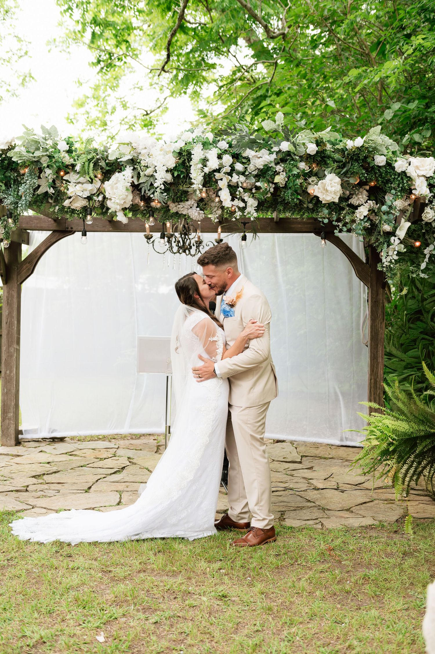 Bride and groom share first kiss under floral arch at outdoor wedding ceremony.