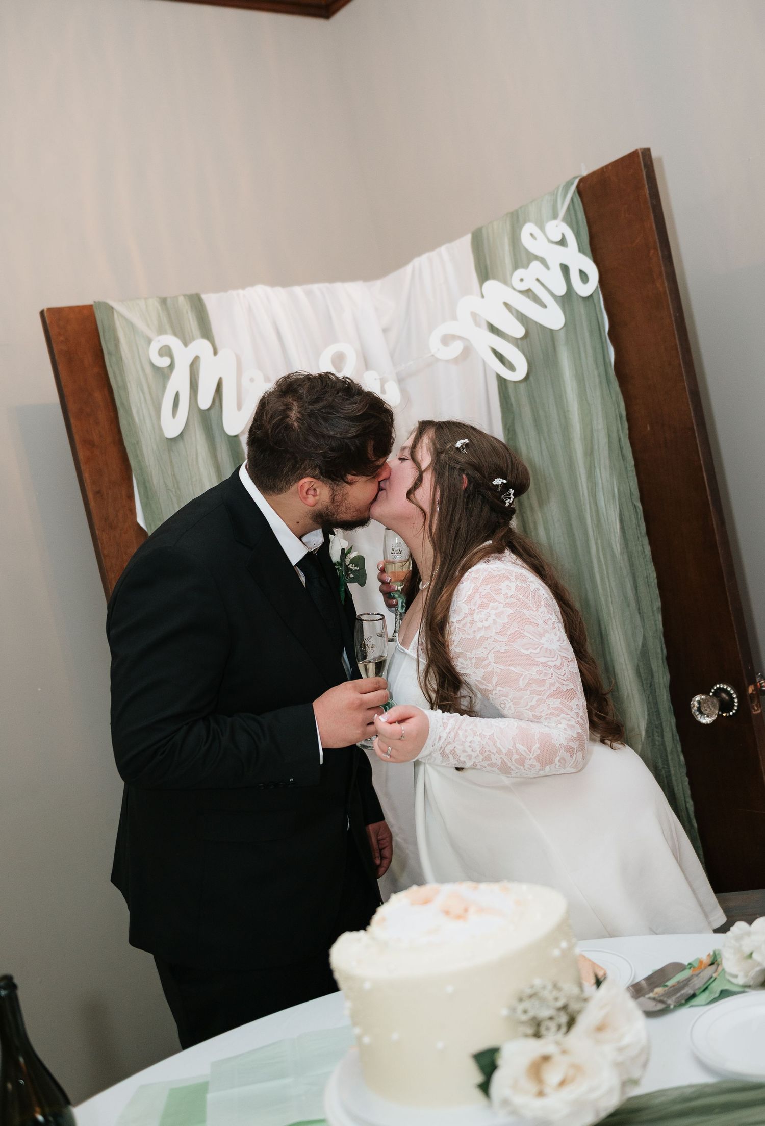 Newlywed couple sharing a kiss at wedding reception with Mr. & Mrs. banner backdrop and white wedding cake in foreground.