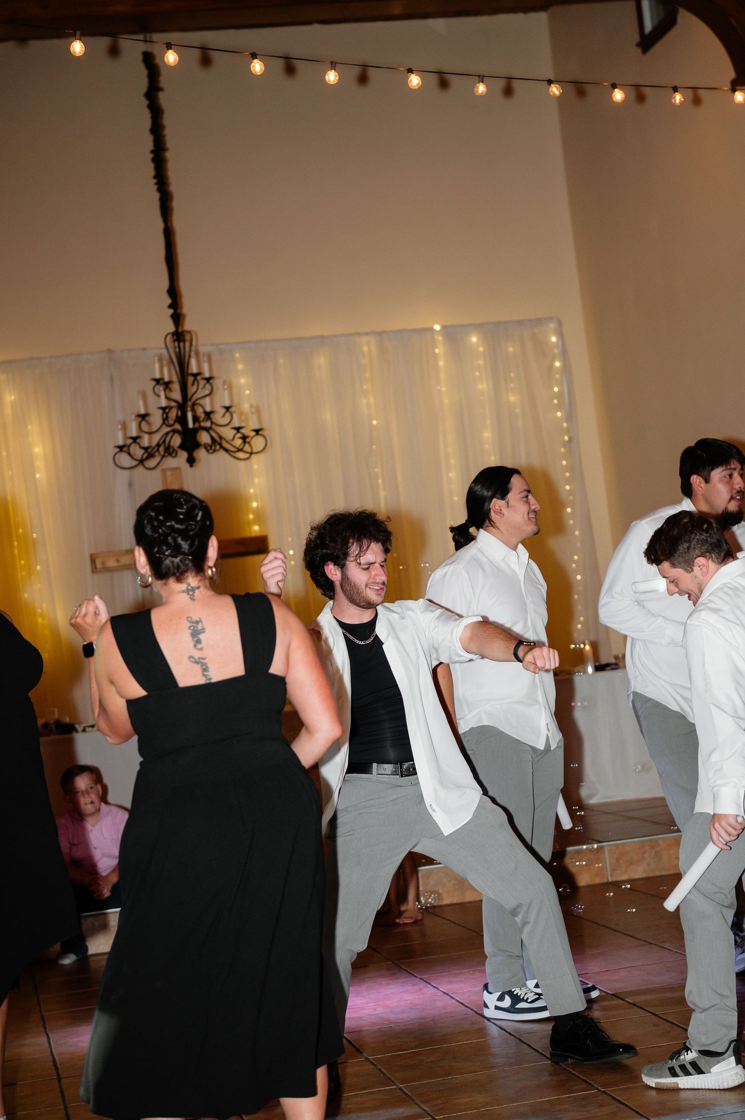 Guests dancing at a wedding reception in a decorated venue with string lights and white drapes.