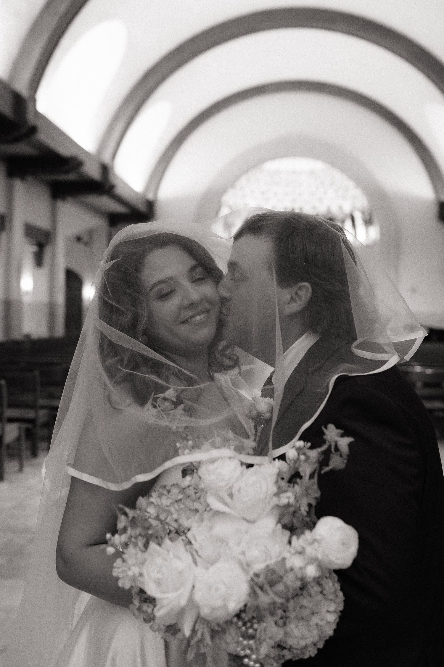 Black and white photo of a smiling bride holding flowers as groom kisses her cheek inside a church.