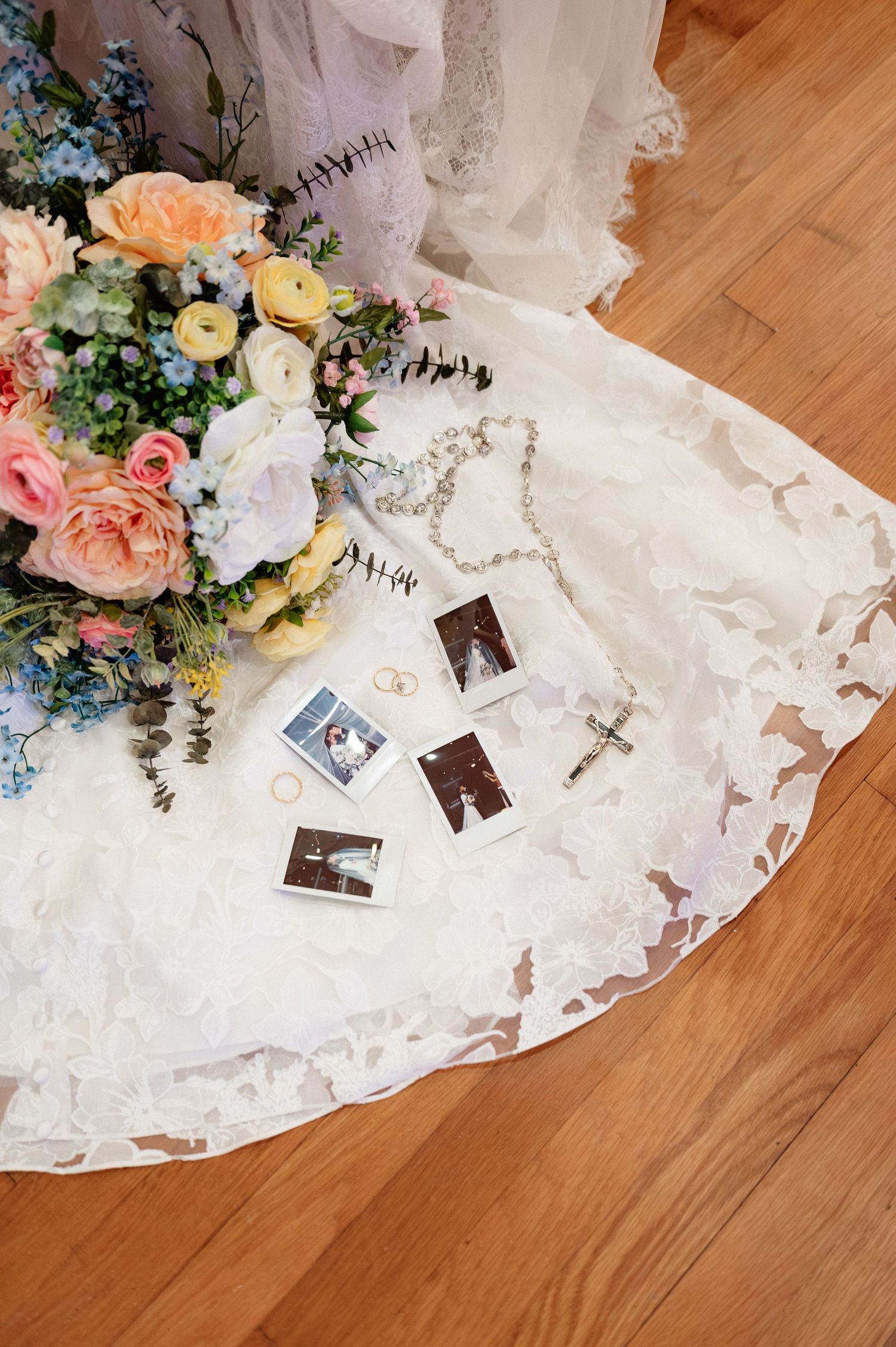 Bridal flat lay with Polaroid photos, pearl rosary, rings, and colorful floral bouquet on white lace wedding dress on wood floor.