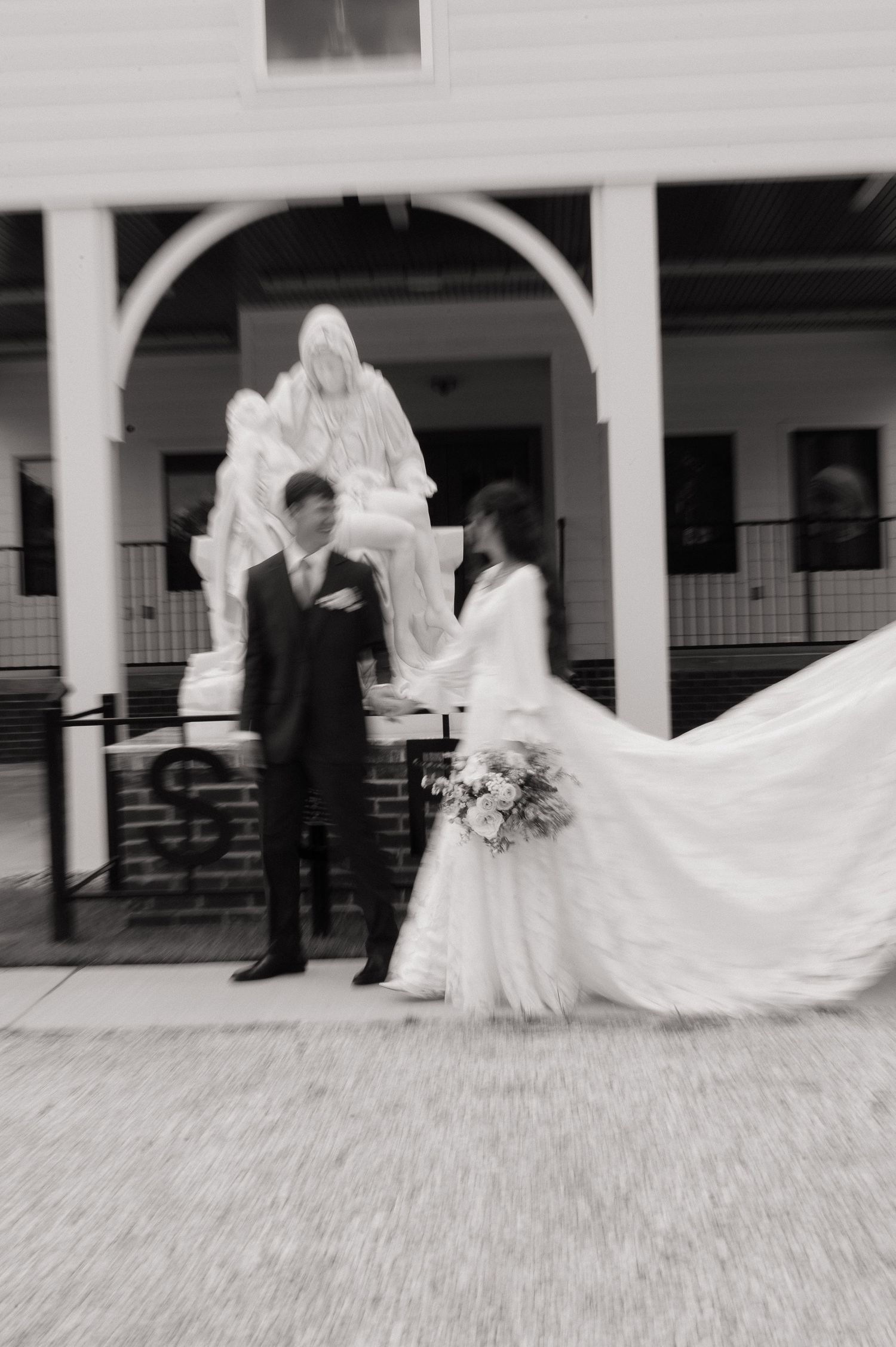 Black and white wedding photo of bride and groom standing before a religious statue outside a church.