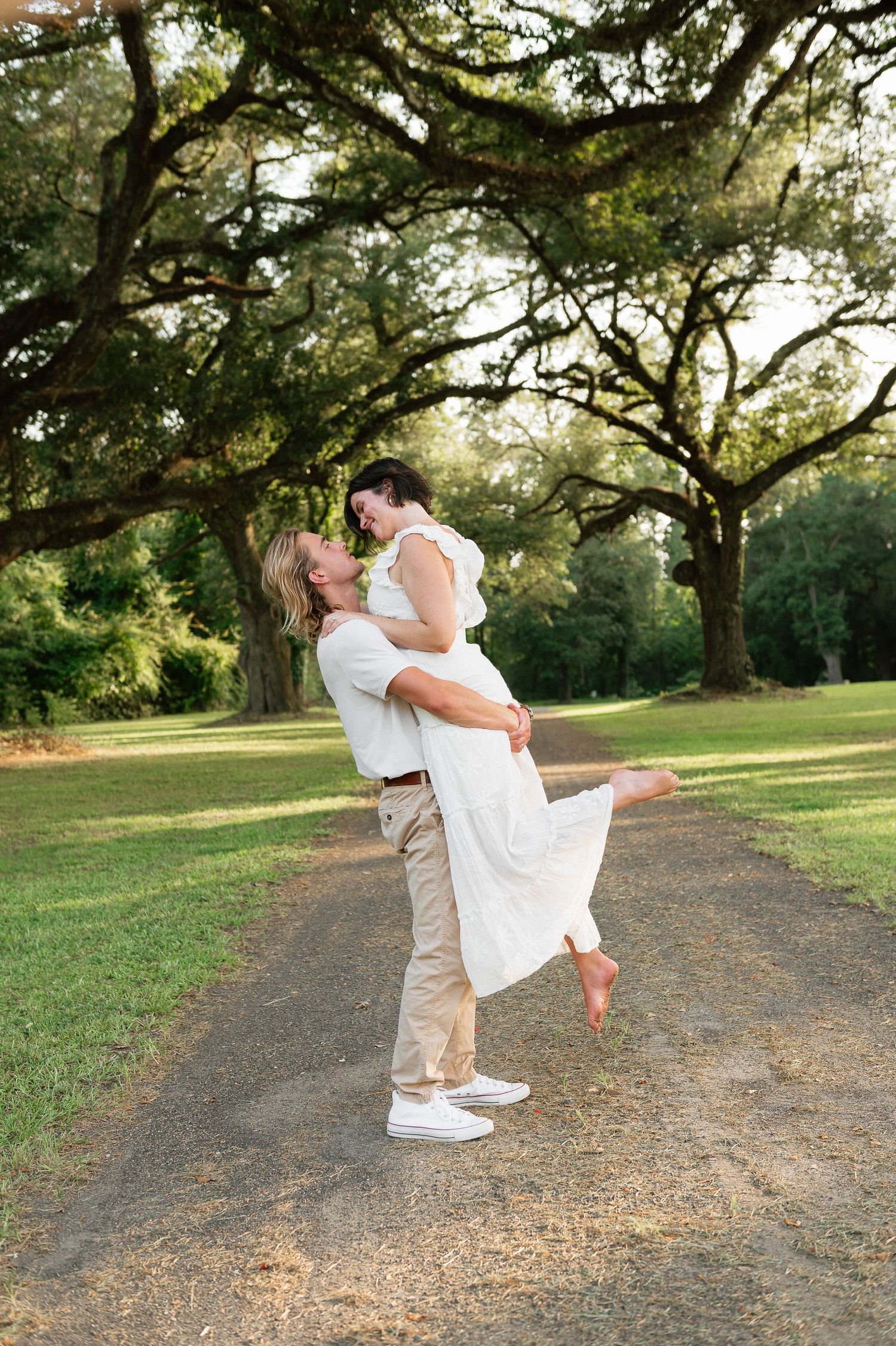 Couple sharing a romantic lift and kiss on a tree-lined path in a lush green park during golden hour.