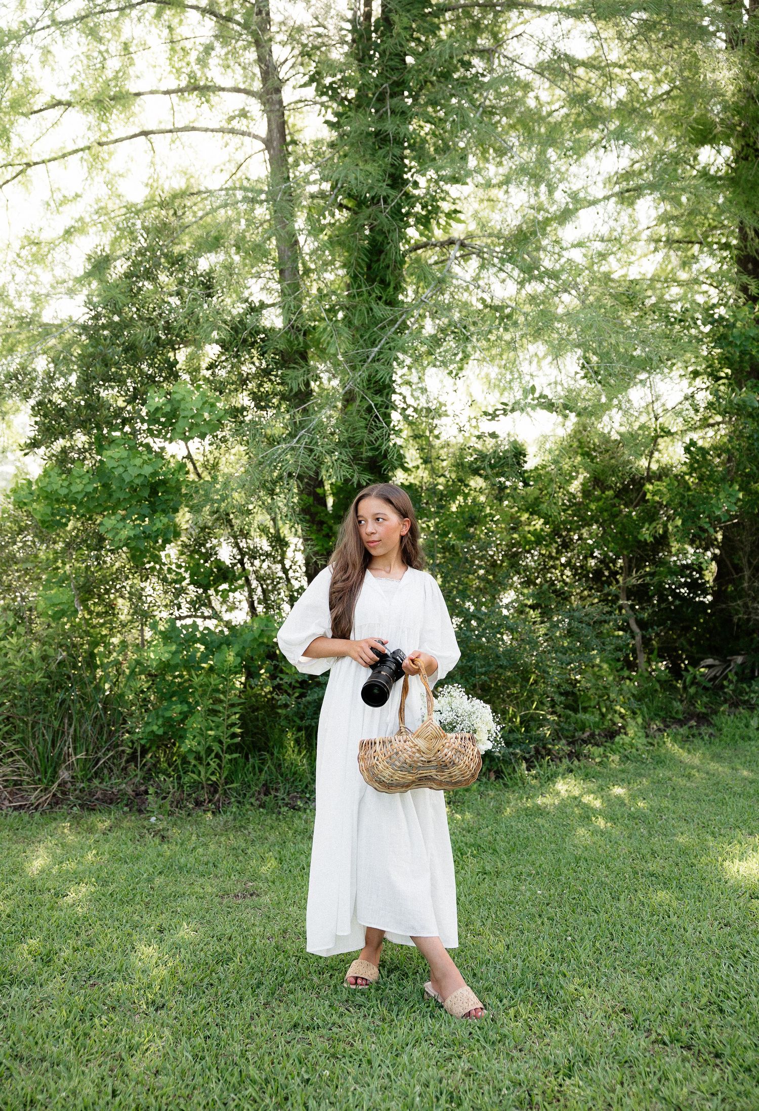 Woman in white dress holding camera and floral basket stands in lush green garden setting.