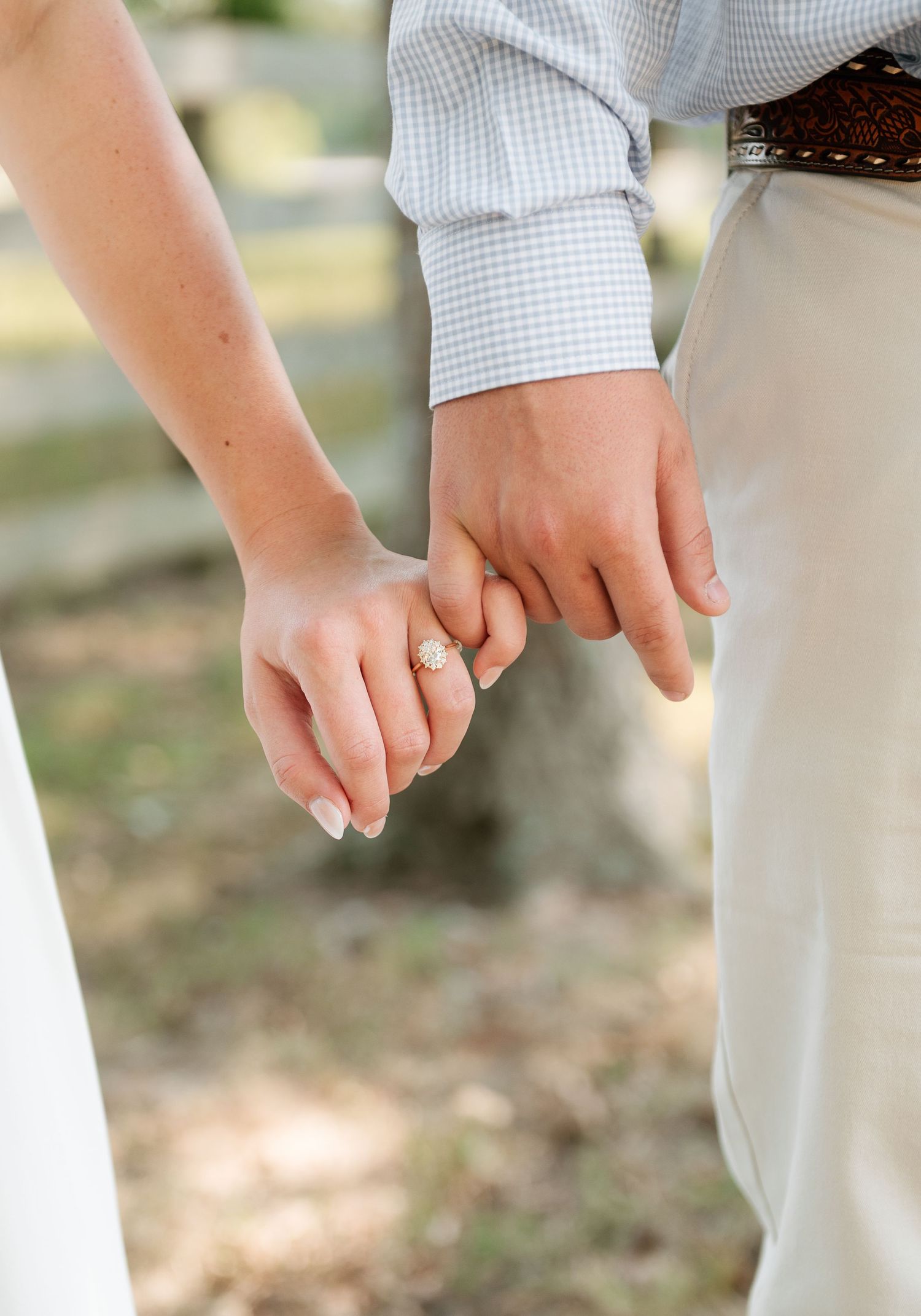Couple holding hands outdoors, with a close-up of her sparkling engagement ring and his checked shirt and leather belt.