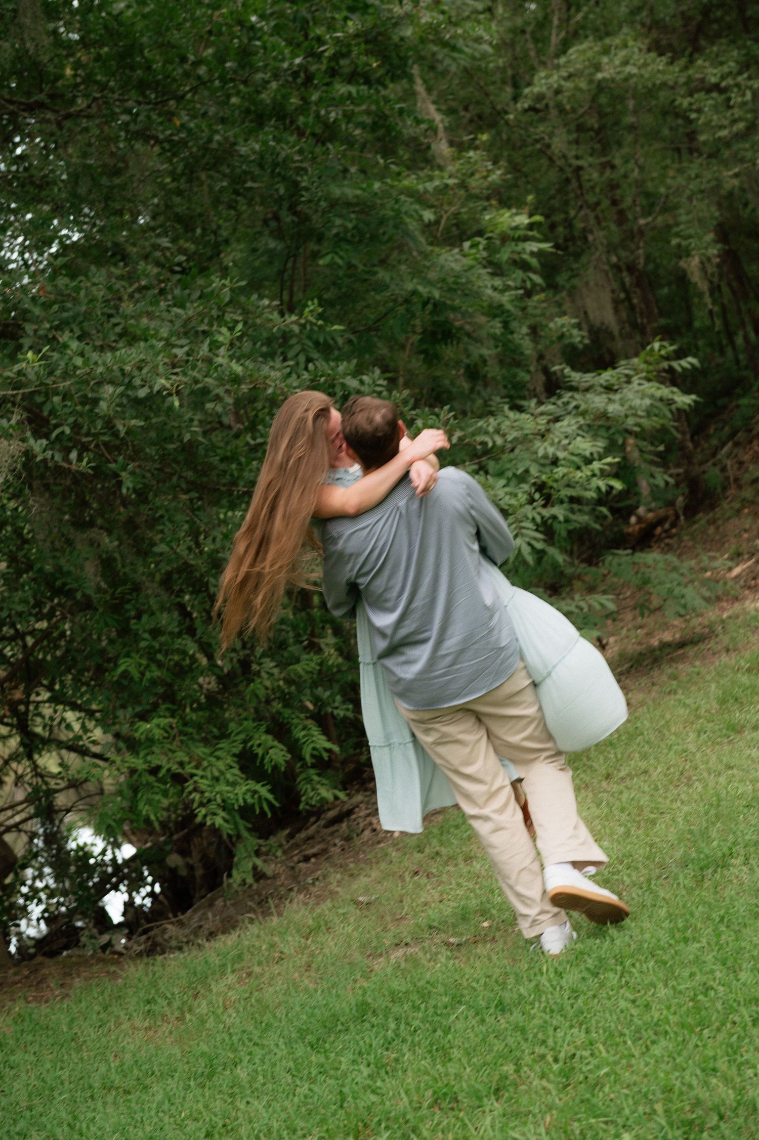 Couple embracing outdoors near lush green trees, man lifting woman in gray dress on grassy lawn.