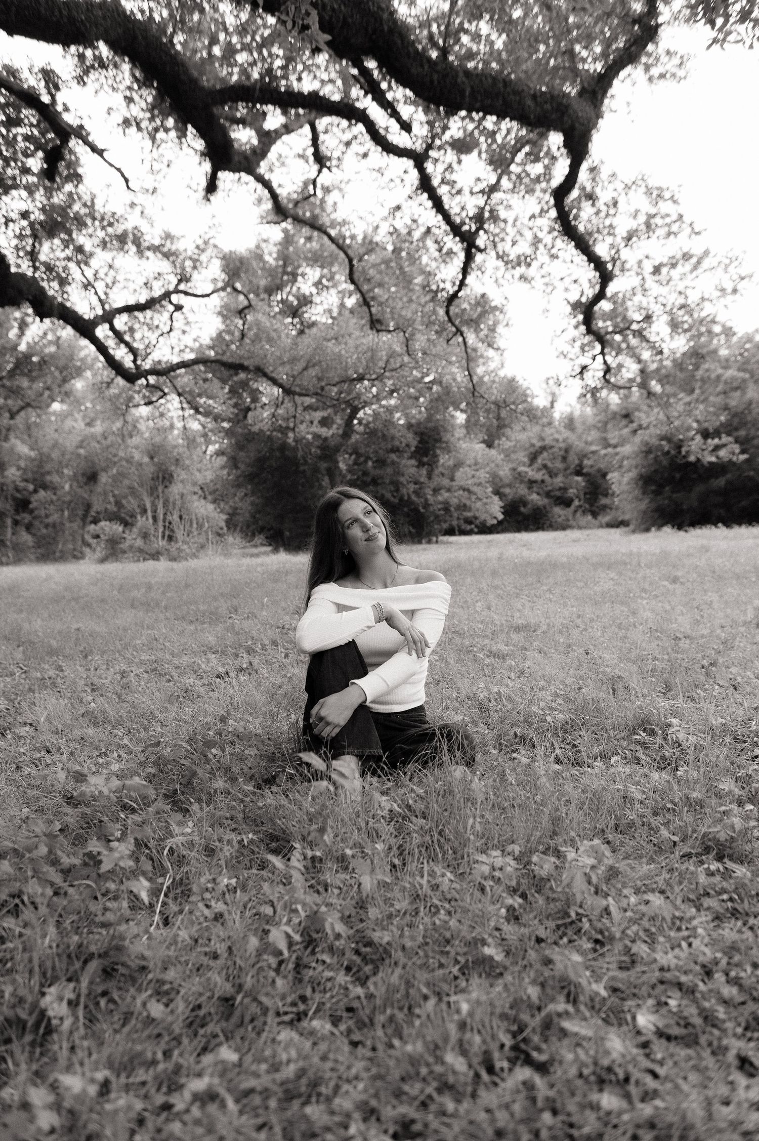 Black and white photo of a young woman sitting in a grassy field, arms crossed, beneath sprawling oak tree branches.