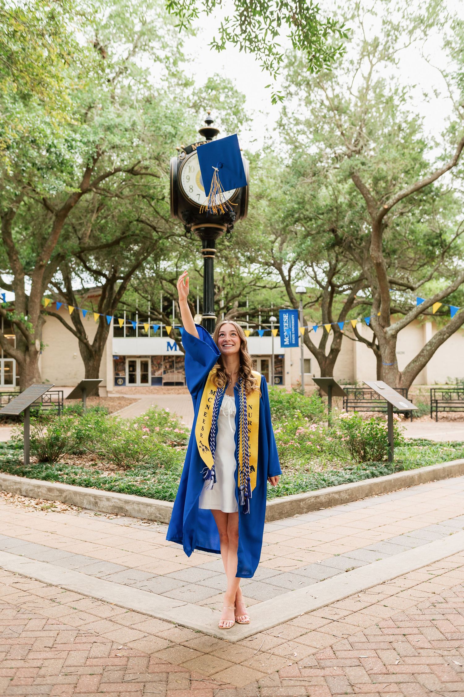 Young woman in blue graduation gown and sash celebrates outdoors on a brick plaza with trees and campus buildings.