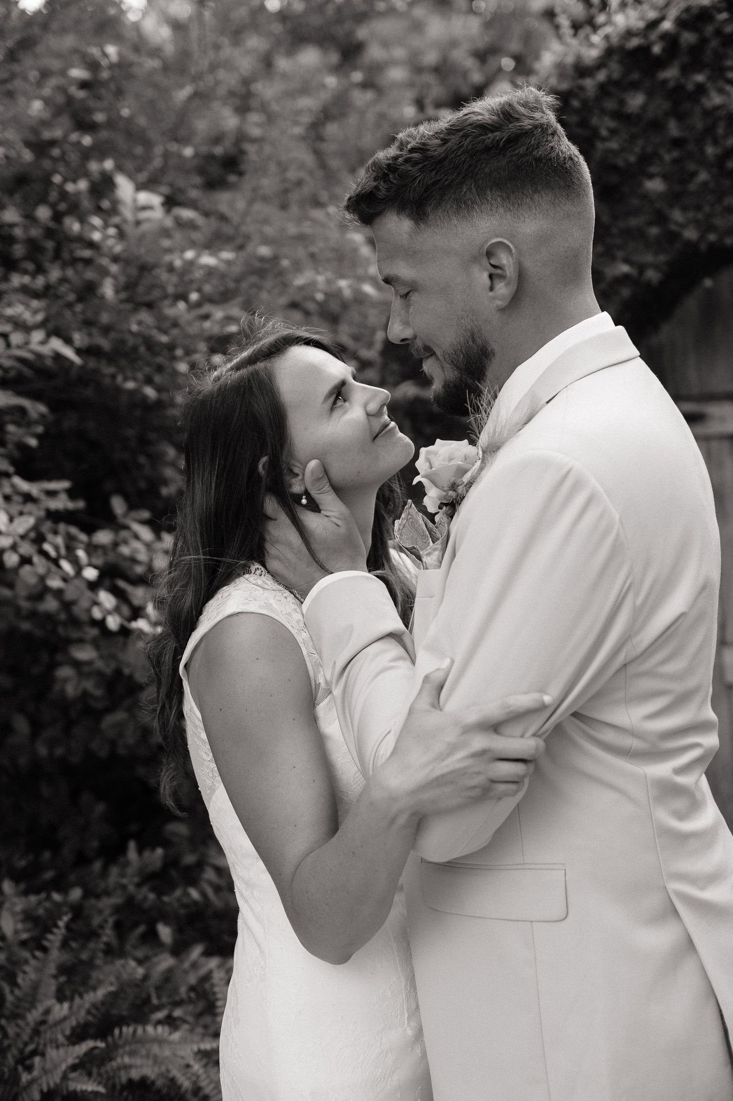 Black and white photo of a couple in wedding attire sharing a romantic moment outdoors among lush greenery.