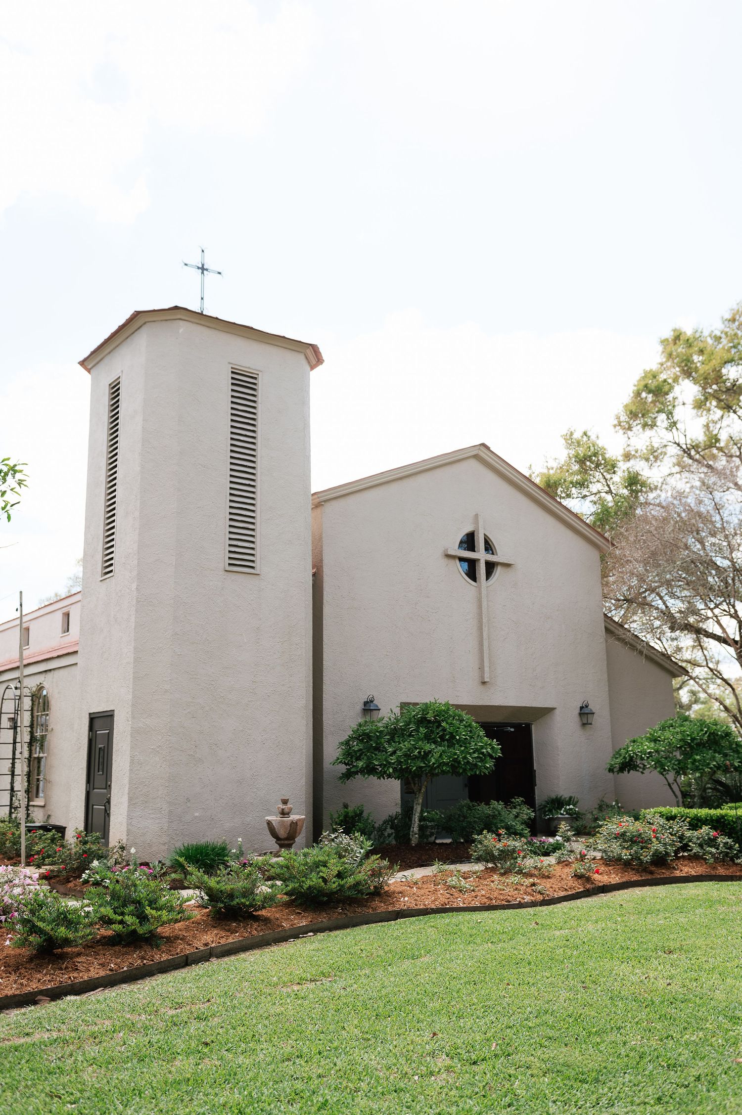 White stucco church with bell tower, cross window, and lush landscaping under a bright sky.