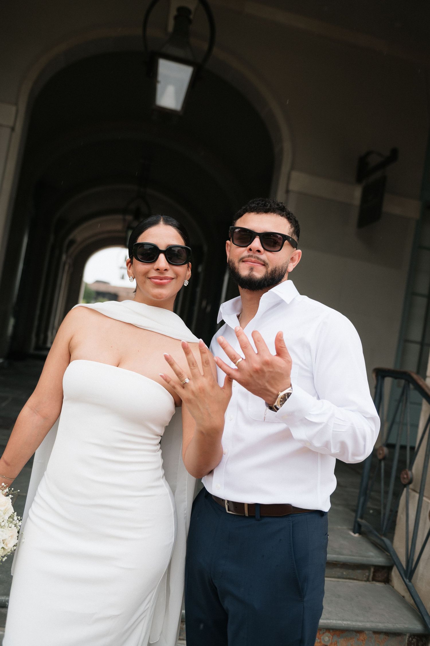 Newlywed couple in sunglasses showing off wedding rings, bride in white gown, groom in white shirt and navy pants.