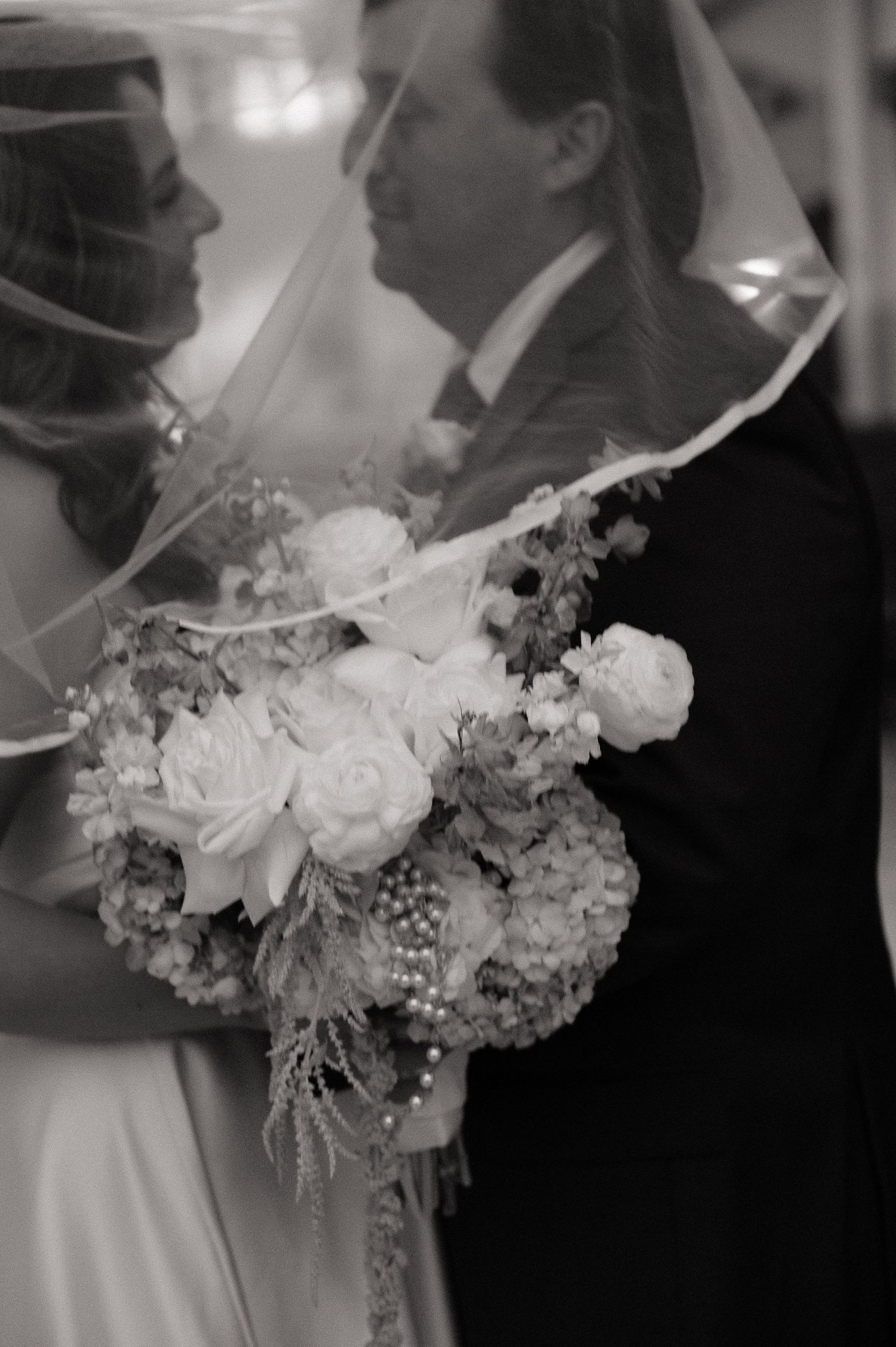 Close-up black and white photo of bride and groom sharing intimate moment under veil with floral bouquet.