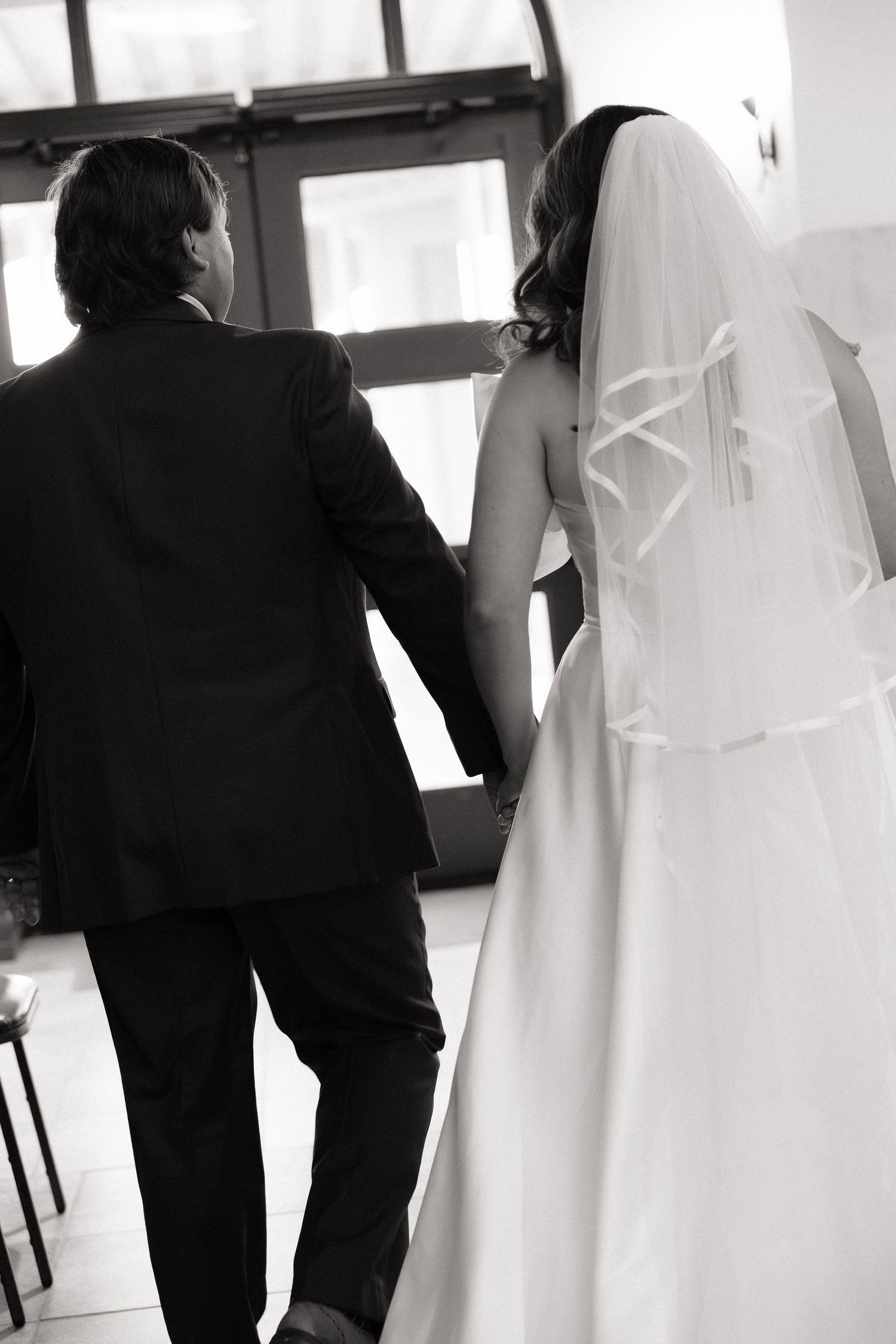 Black and white photo of bride in veil and groom in suit walking hand in hand at wedding.