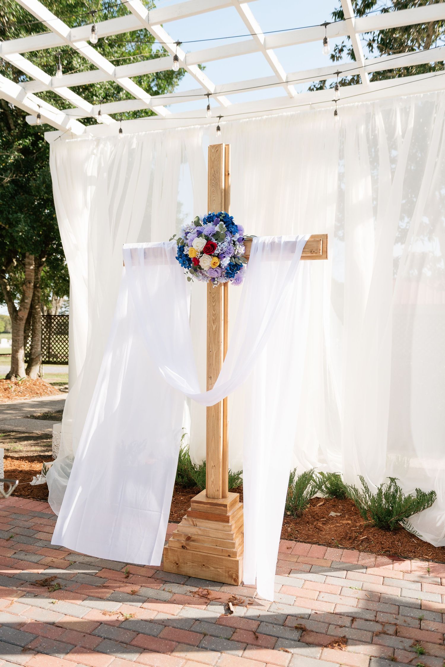 Wooden cross adorned with colorful floral bouquet and white draping fabric at an outdoor wedding ceremony.