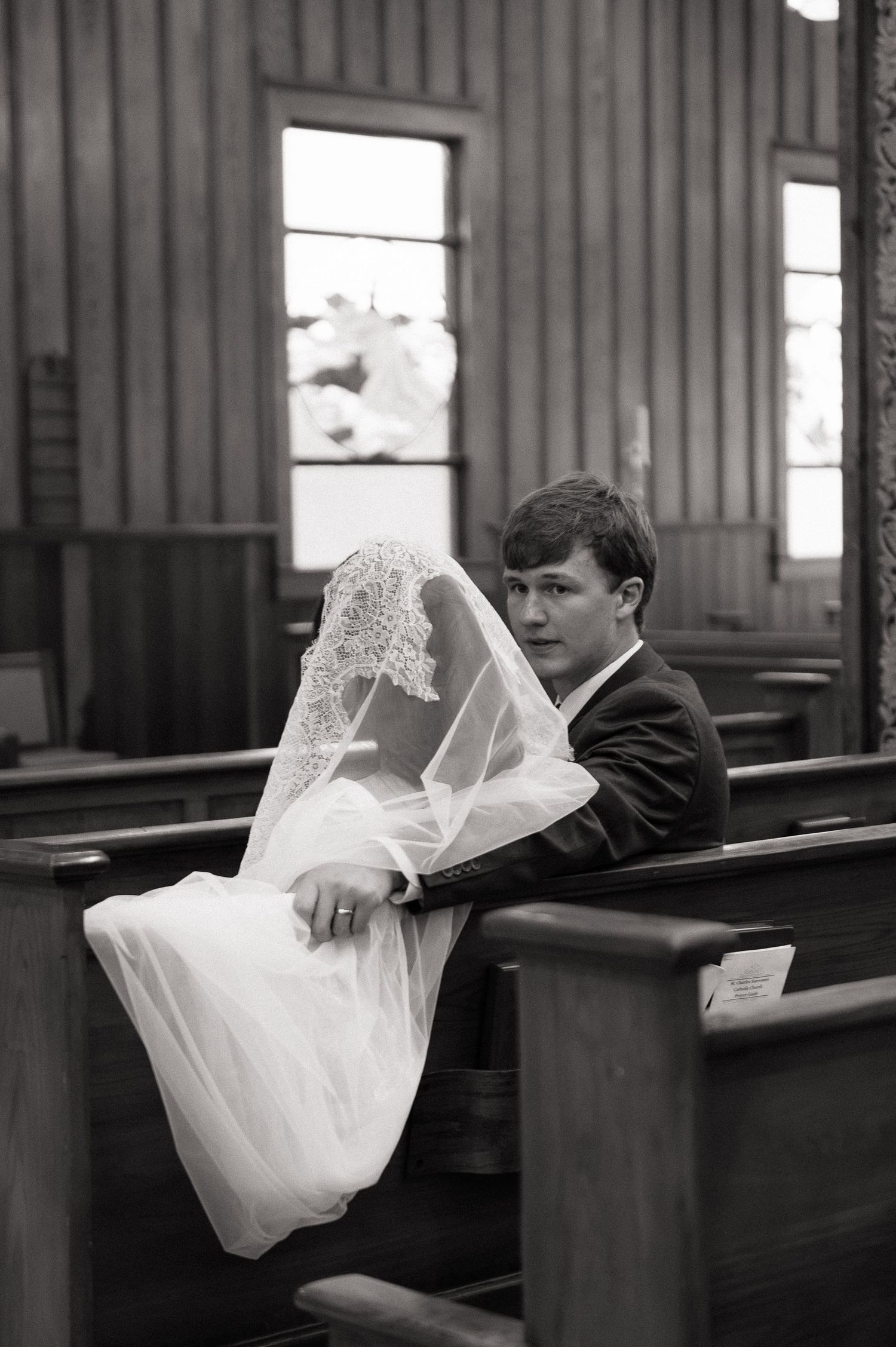 Black and white photo of a groom in a suit sitting in church pews beside a bride in a white gown and veil.