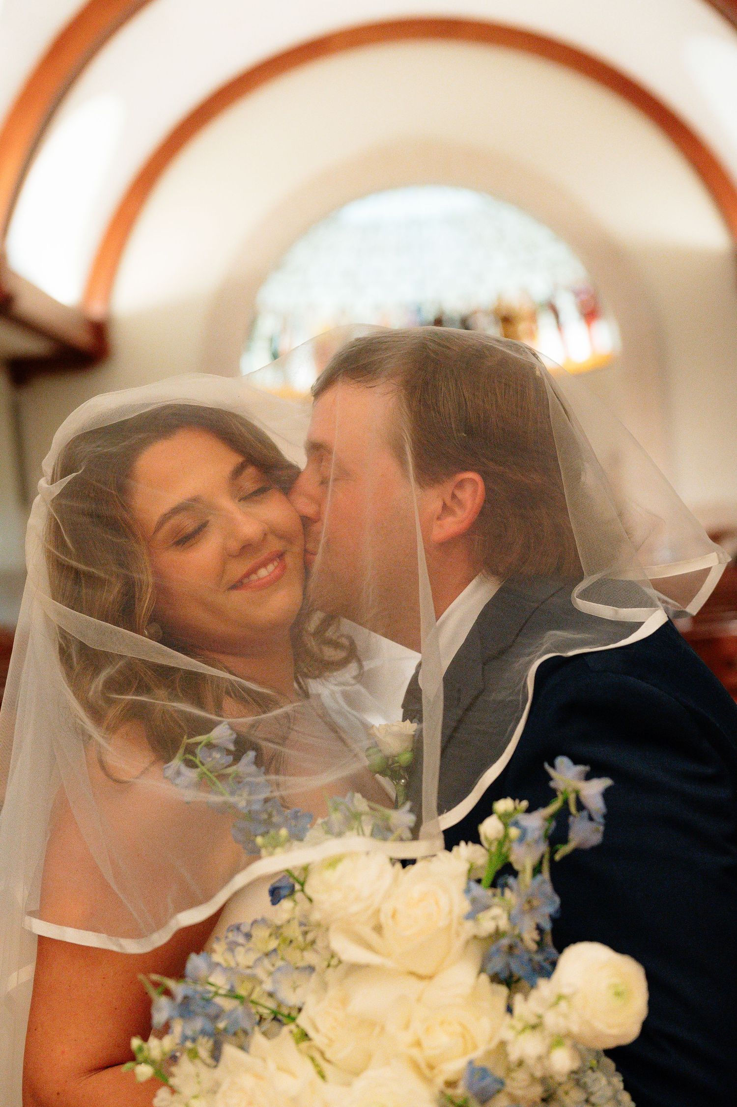 Groom kissing bride under veil in church as she holds blue and white wedding bouquet.