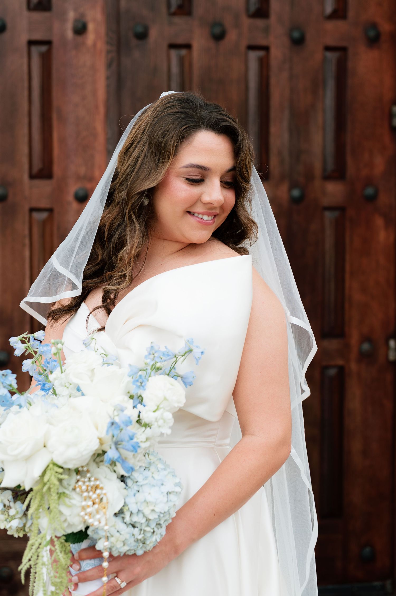 Smiling bride in white gown and veil holds blue and white hydrangea bouquet before wooden doors.