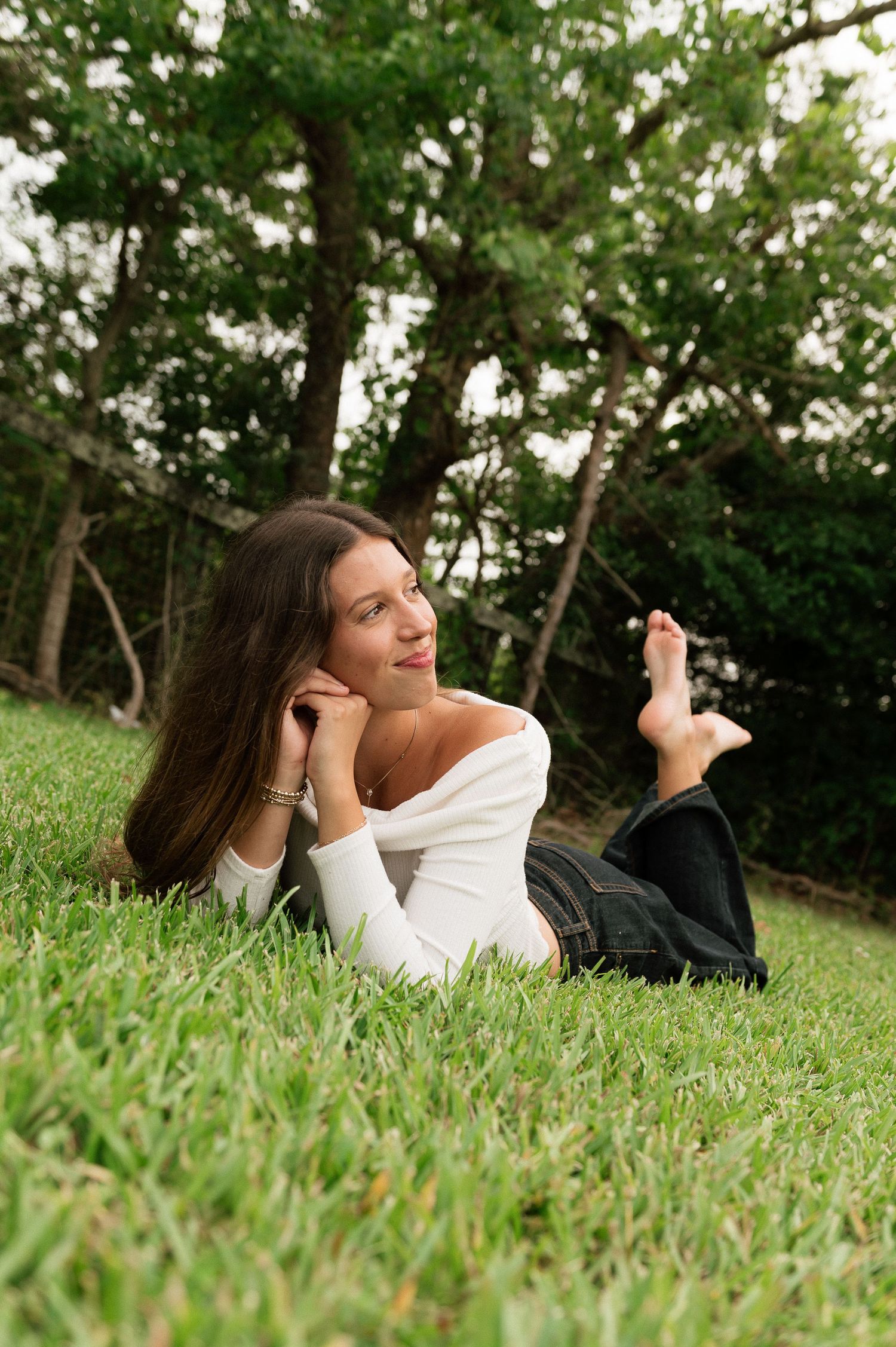 Young woman in white off-shoulder top lying on green grass, gazing thoughtfully, with lush trees in background.
