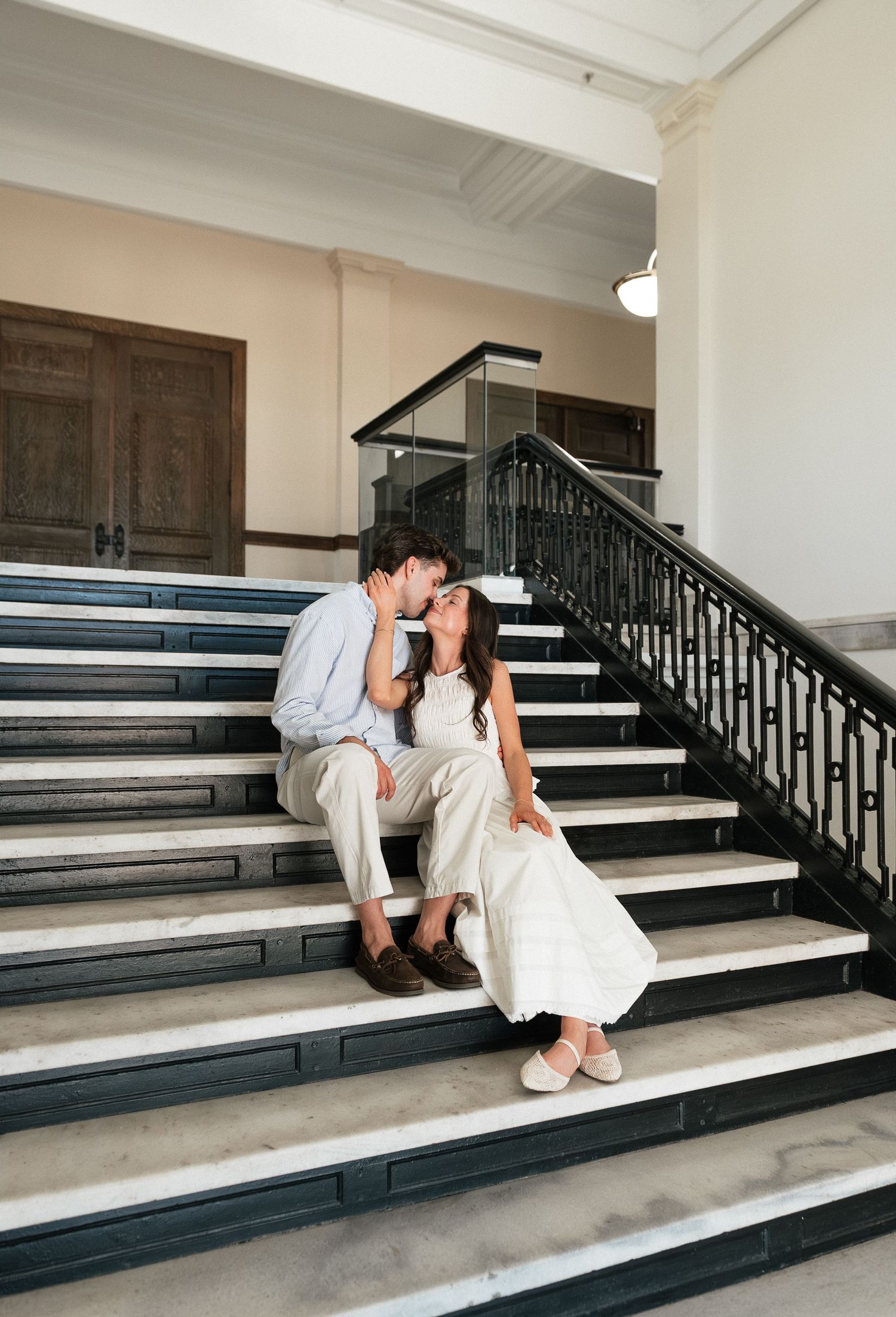 A couple in wedding attire shares a kiss while sitting on elegant marble stairs with black iron railings.