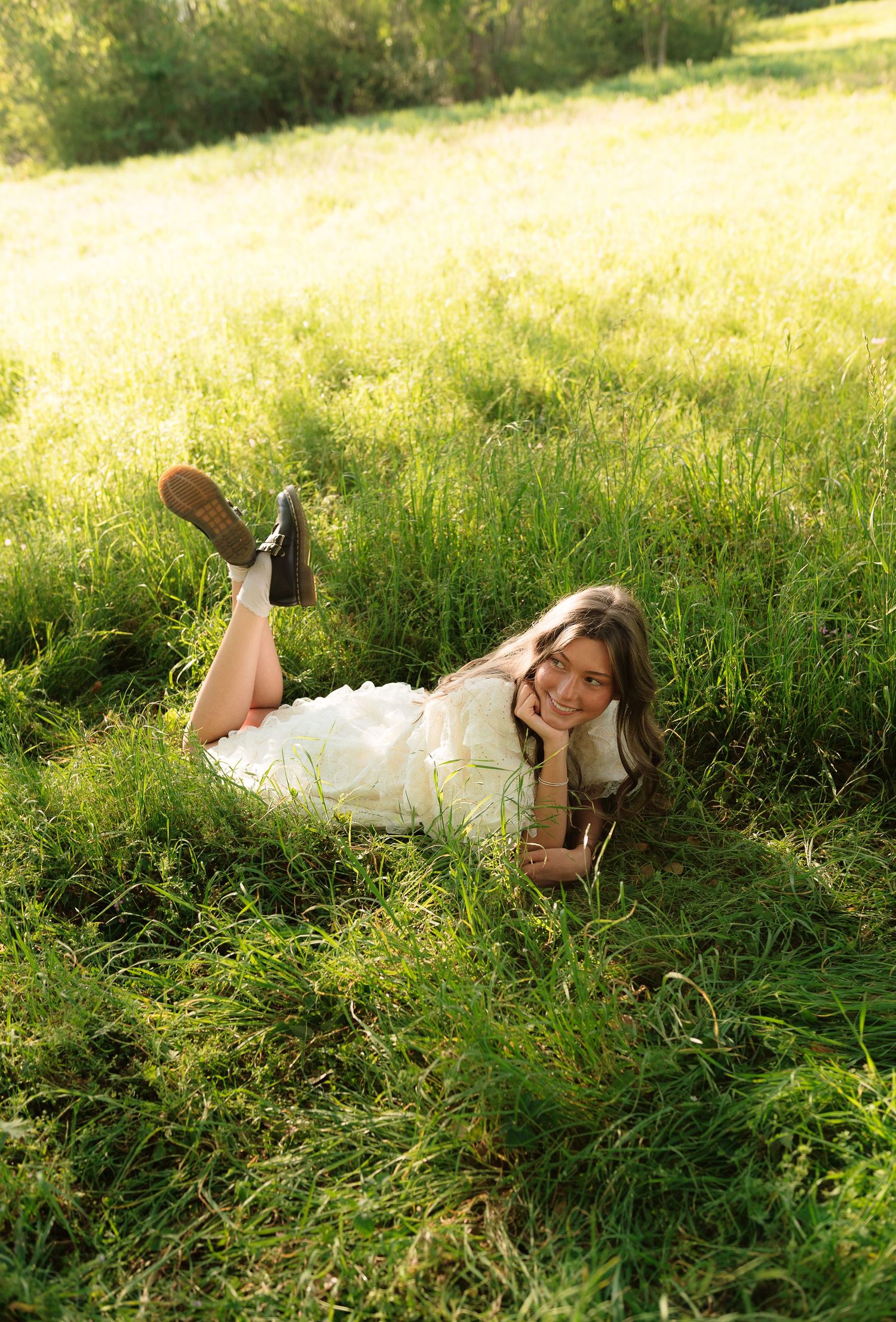Young woman in white dress and boots lying on lush green grass field, smiling in golden sunlight.