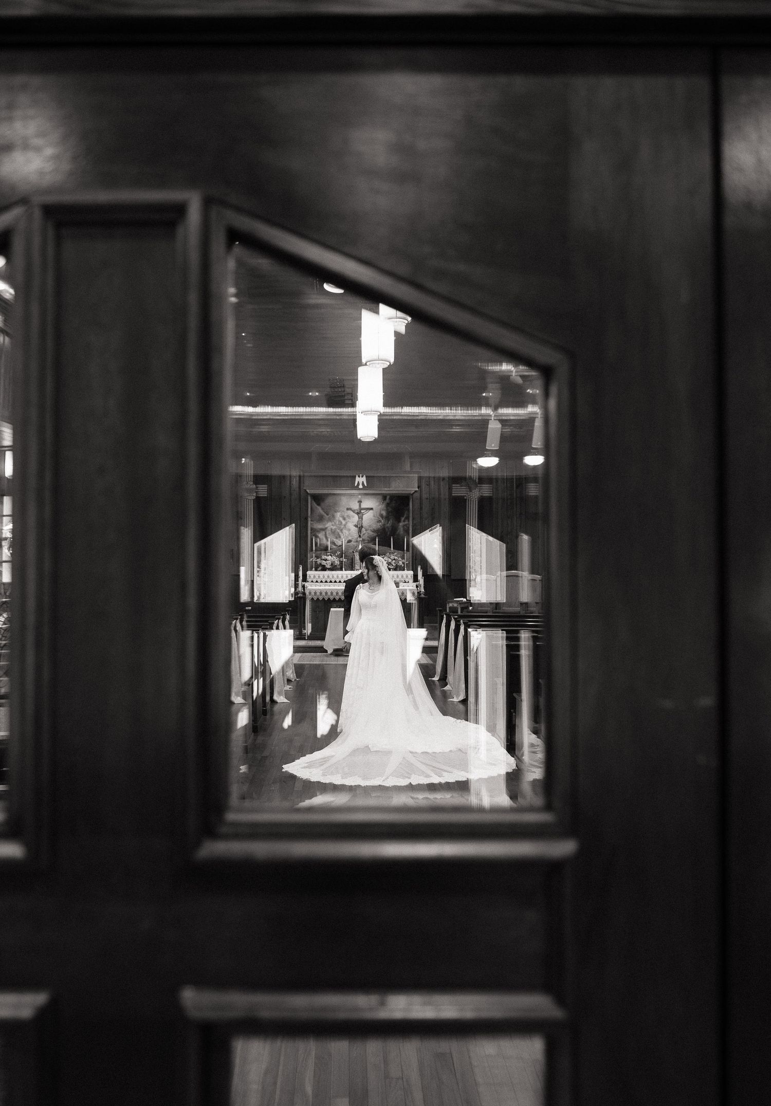 Black and white photo of a bride in a flowing gown and veil standing in a church aisle, viewed through wooden doors.