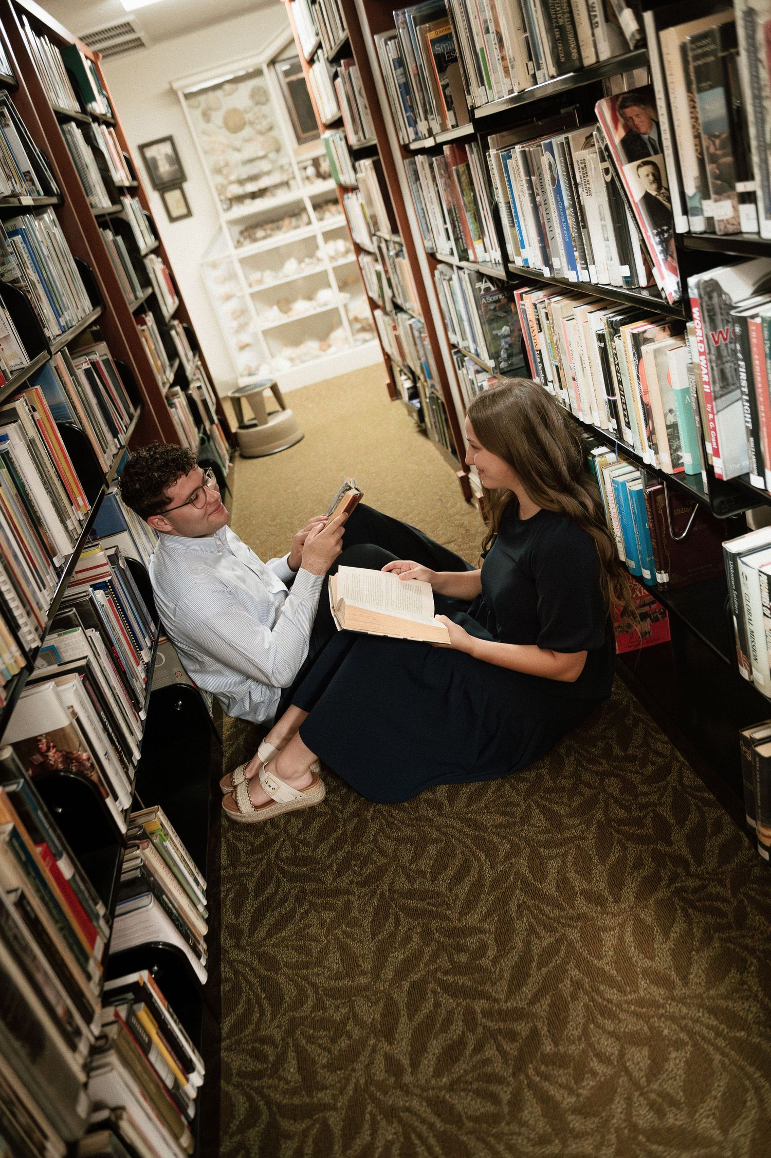 Two people sitting on a library floor between bookshelves, reading and sharing books together.
