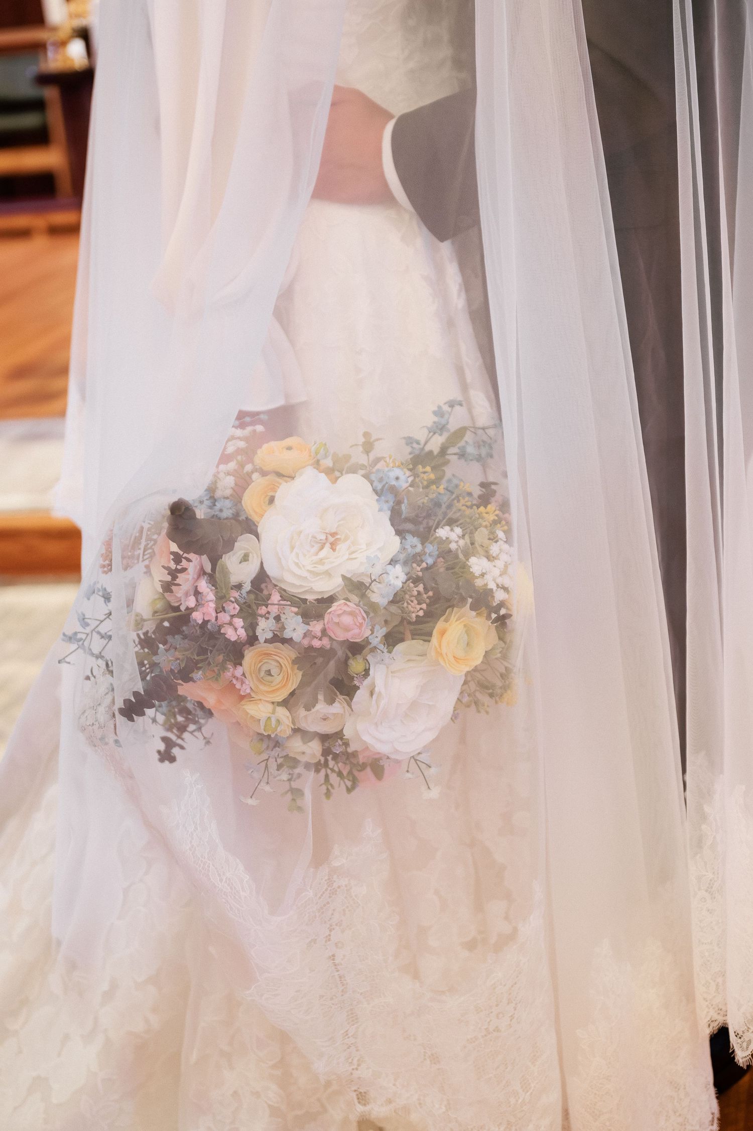 Bride in lace wedding gown holding a pastel bouquet of white, yellow, and pink flowers draped under a sheer veil.