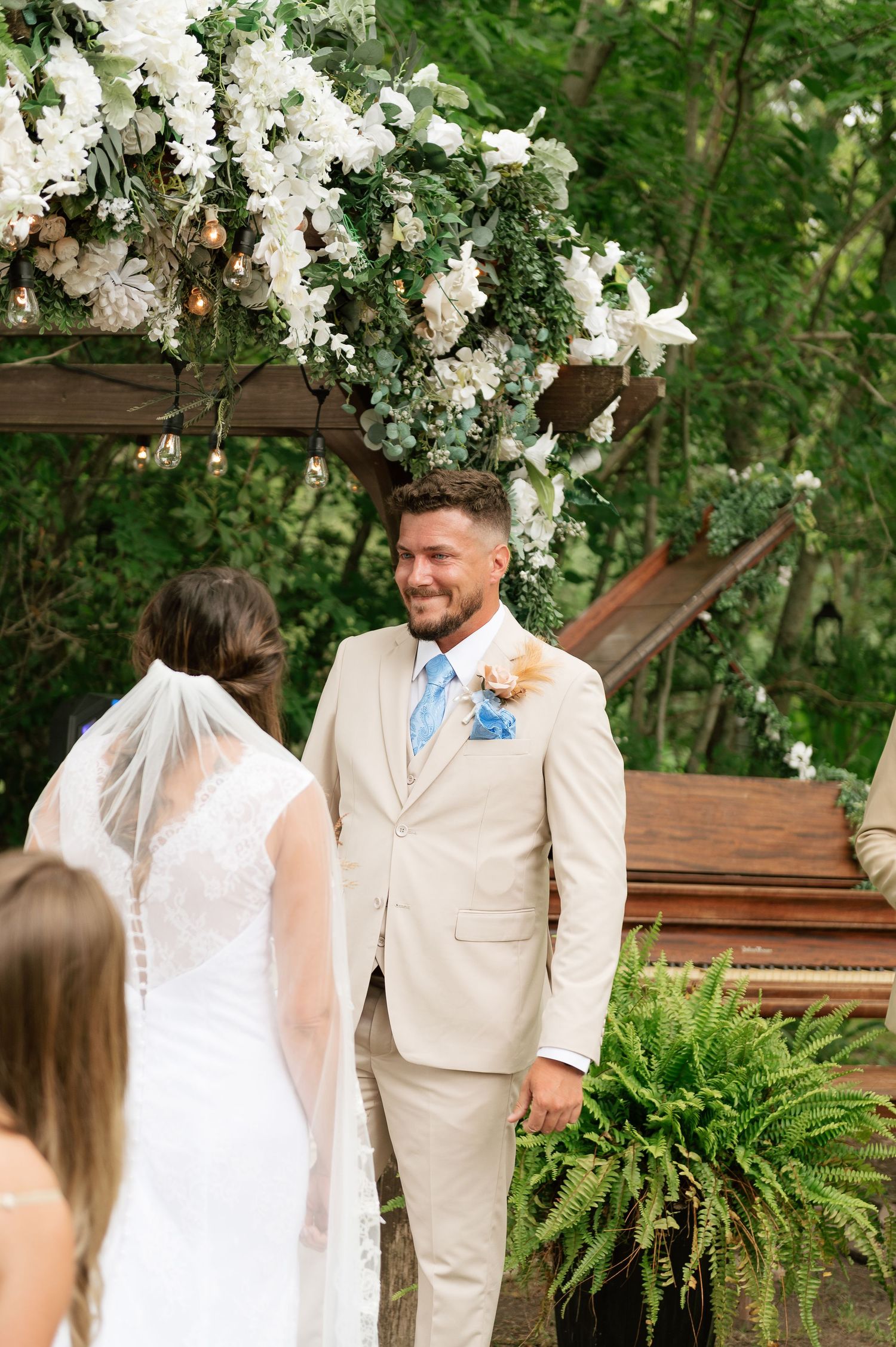 A smiling groom in a tan suit with blue boutonniere stands at the altar facing his bride during an outdoor wedding.