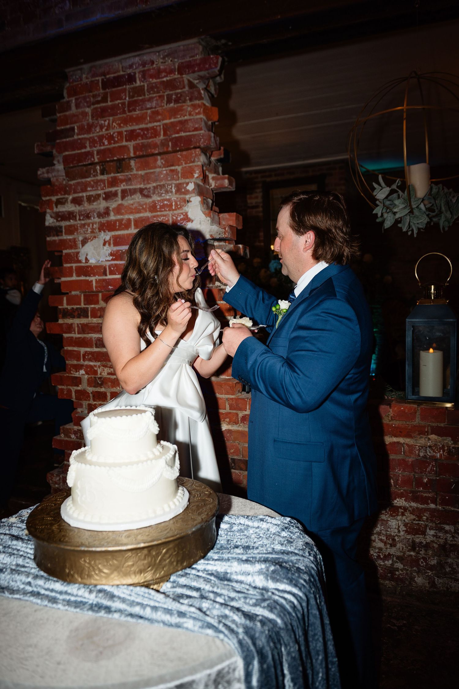Bride and groom feeding each other wedding cake at rustic brick venue reception.