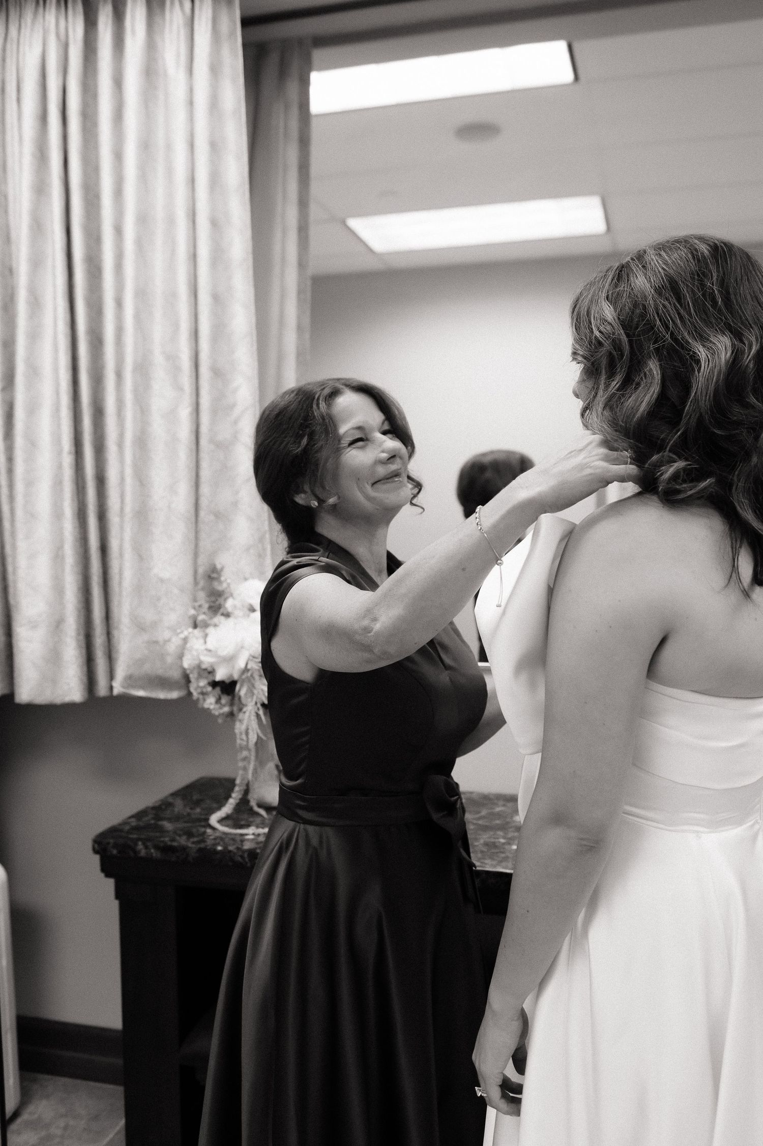 Woman in black dress helps bride with necklace in black and white wedding preparation photo.
