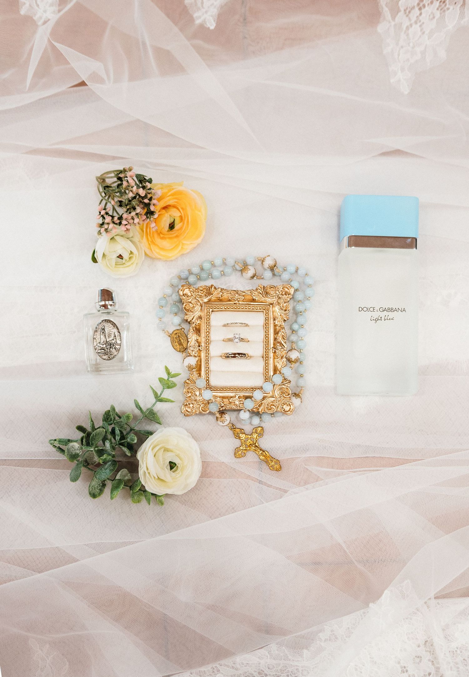 Elegant wedding flat lay with gold rings, rosary, Dolce Gabbana perfume, yellow ranunculus, and lace veil on blush background.