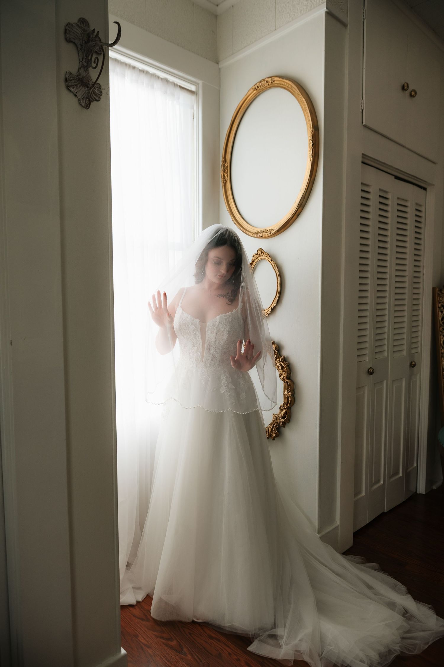 A bride in a flowing white wedding gown and veil stands by a window with ornate gold mirrors on the wall.