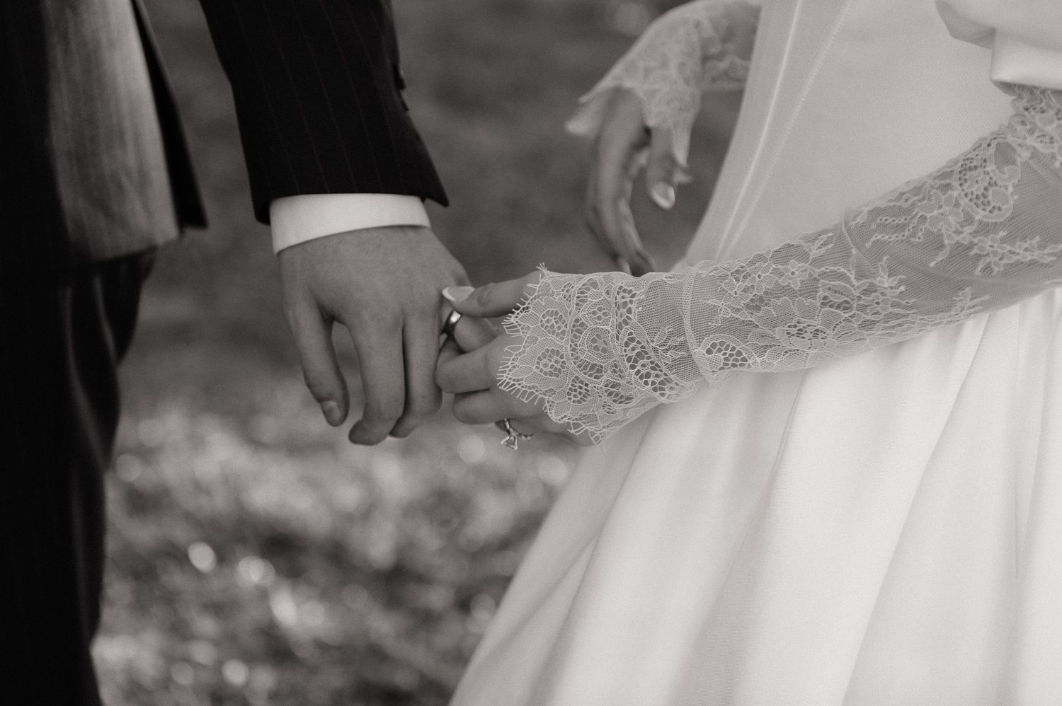 Black and white close-up of newlyweds holding hands, showing lace dress detail.