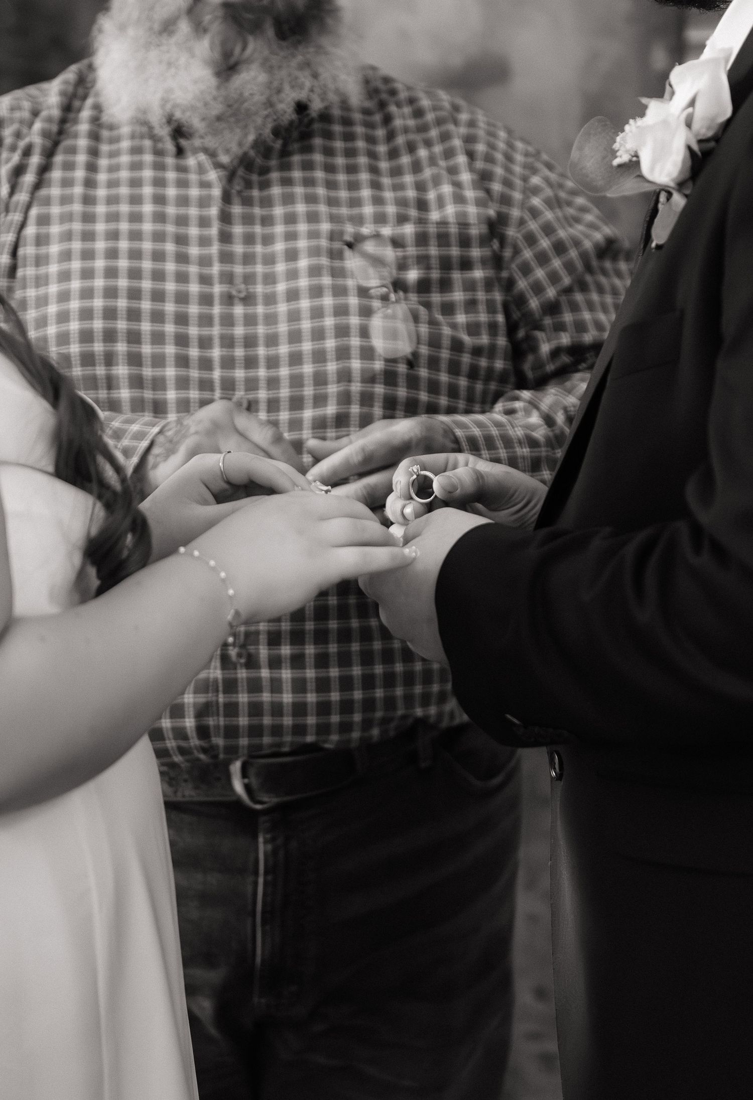 Black and white photo of hands exchanging wedding rings during a ceremony with a plaid-shirted officiant in view.