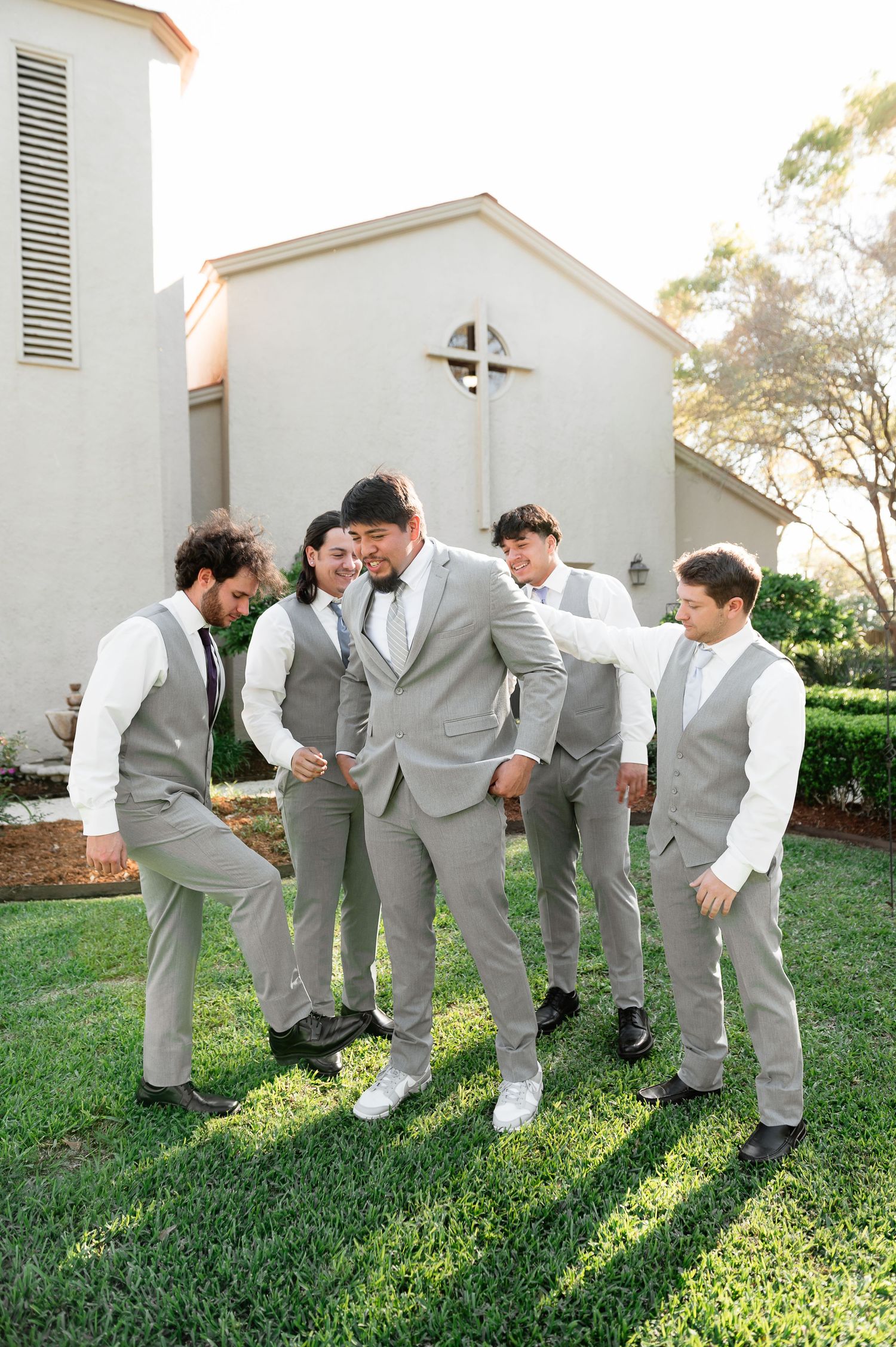 Groom and groomsmen in gray vests pose playfully on green lawn outside white church on wedding day.