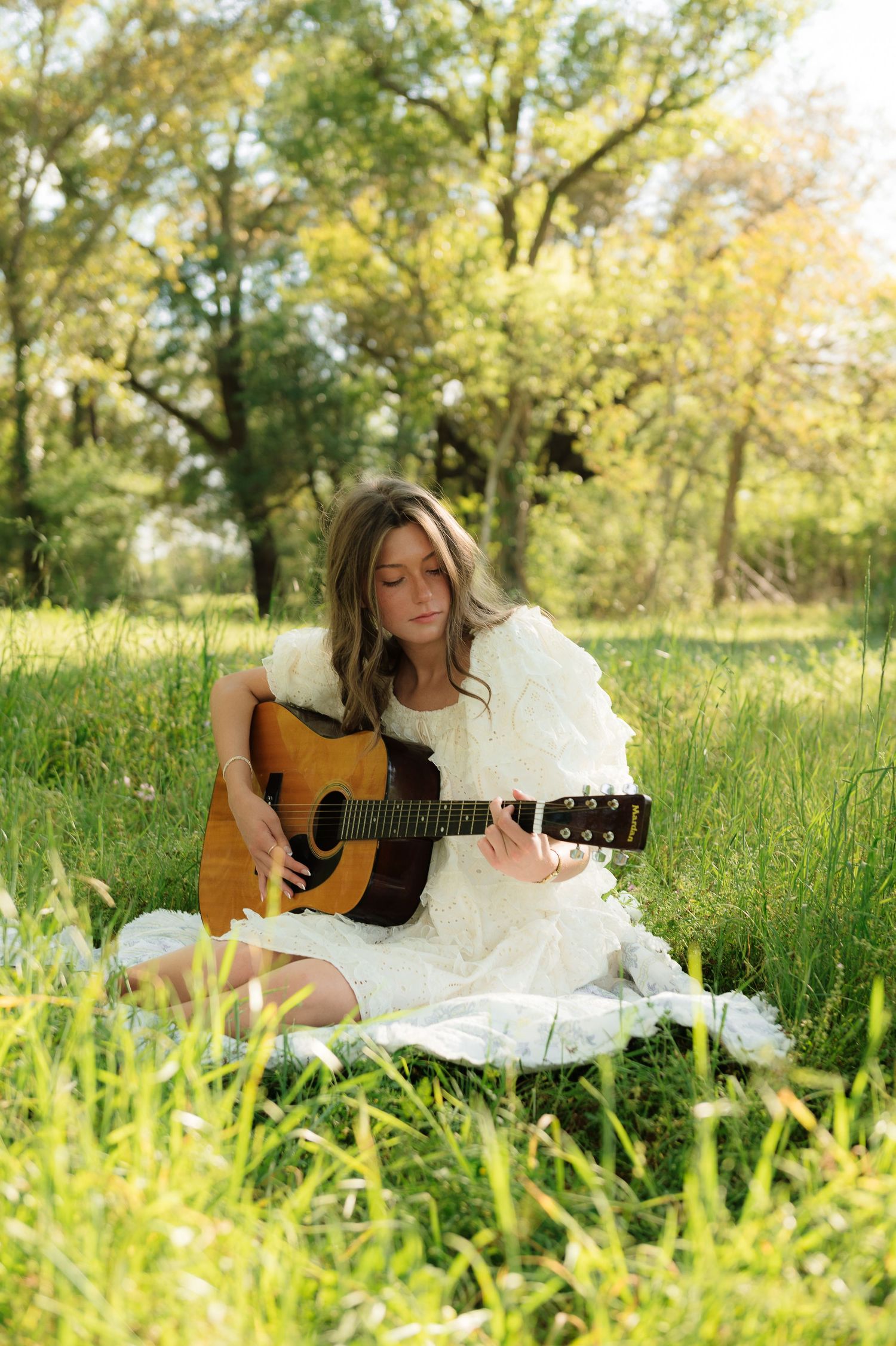 Young woman in white dress playing acoustic guitar while sitting on blanket in sunny green meadow.