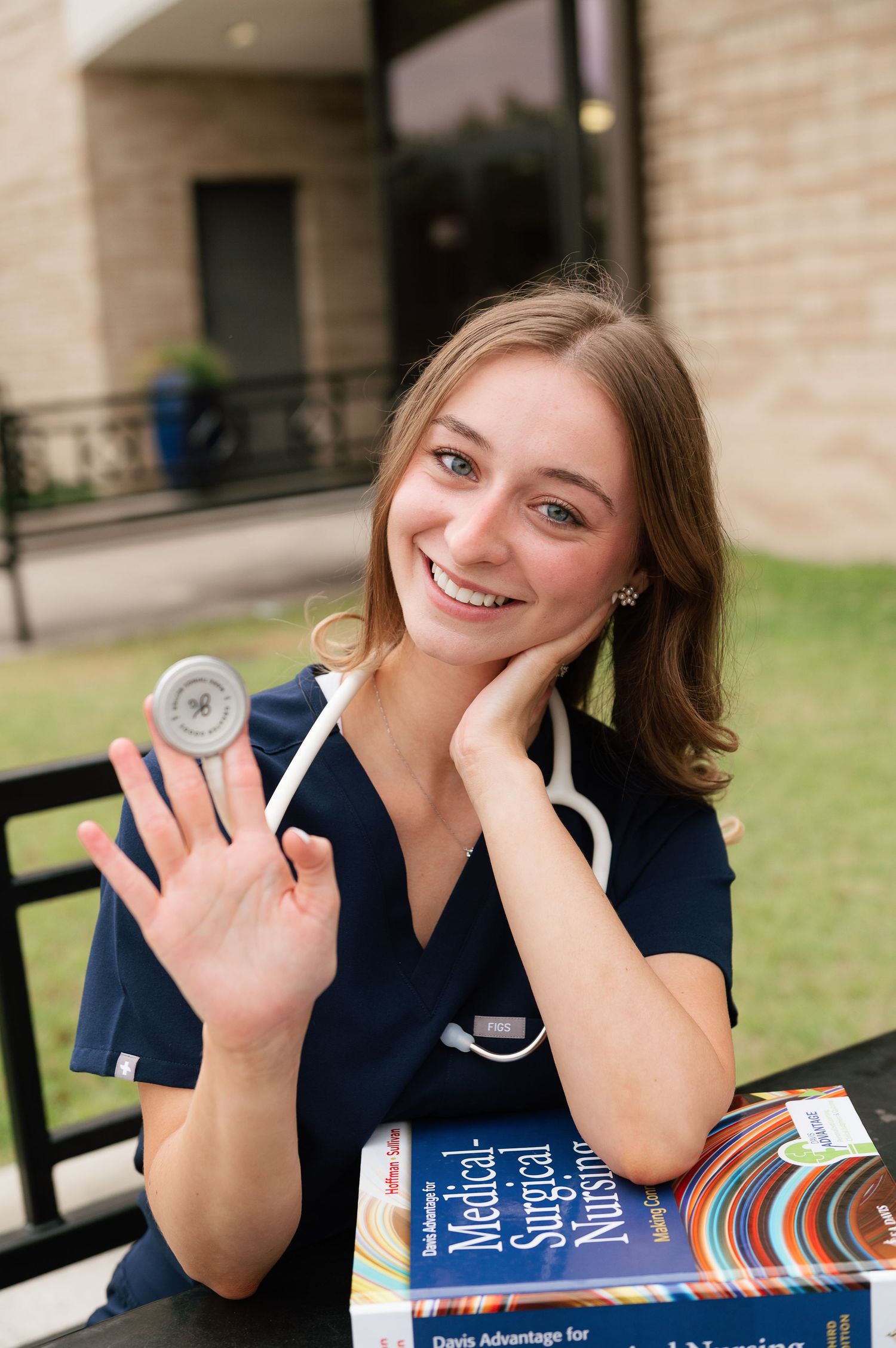 Smiling nursing student in scrubs holding a stethoscope and Medical-Surgical Nursing textbook outdoors.