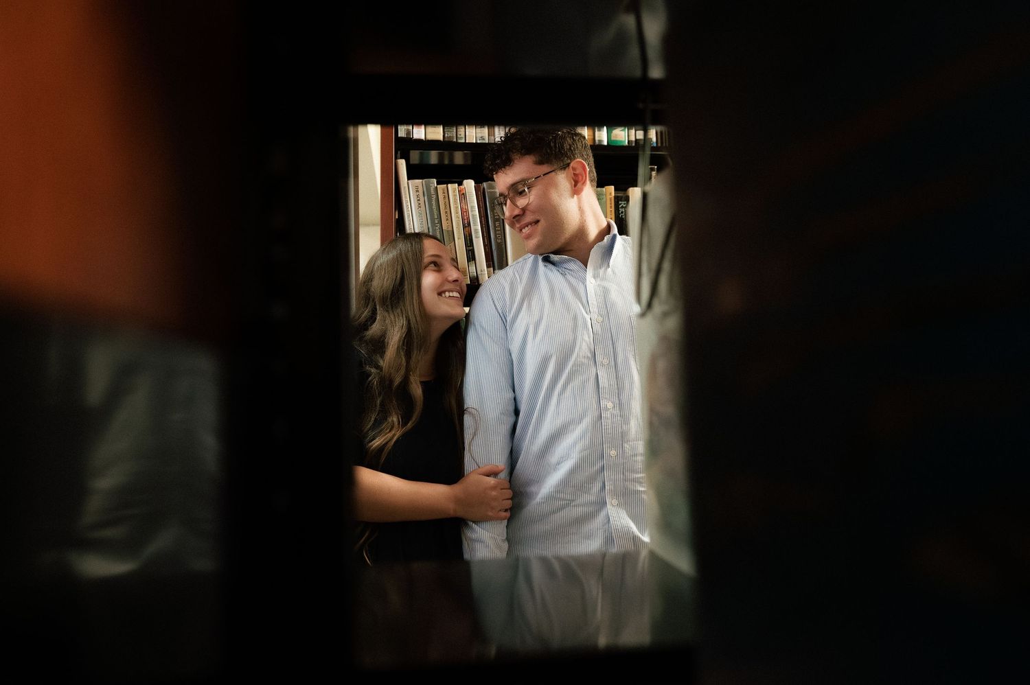 A couple shares a romantic moment smiling at each other between library bookshelves.