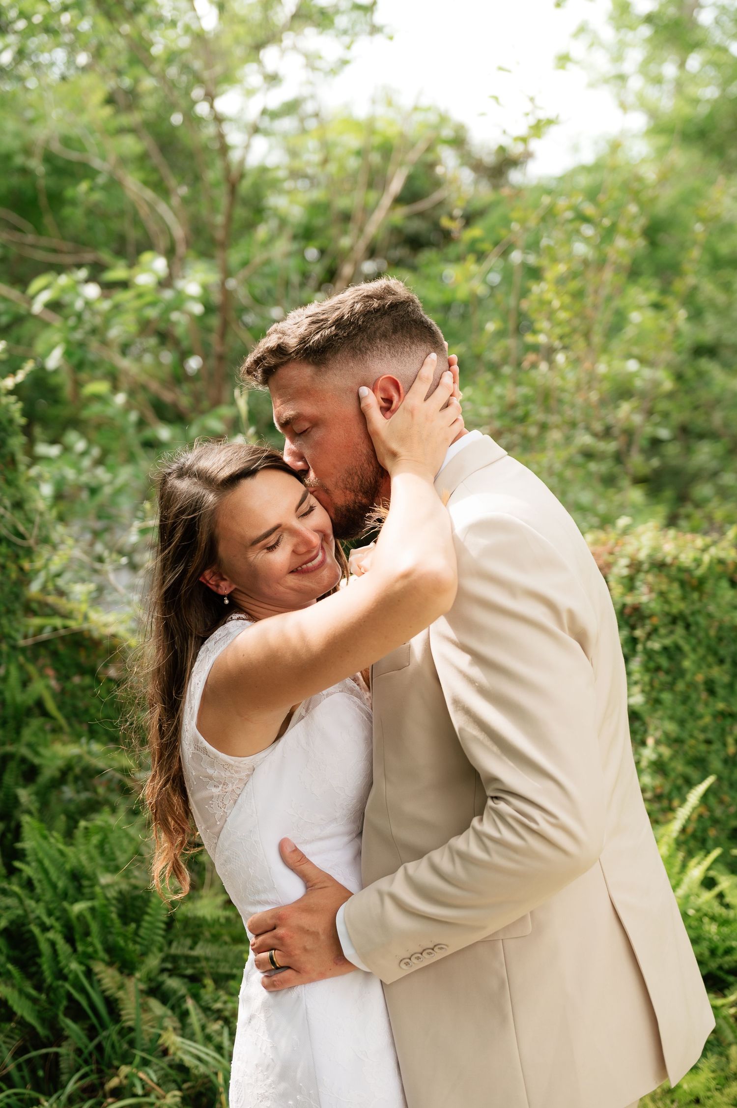 Groom in beige suit kisses bride on cheek while embracing outdoors in green garden wedding portrait.