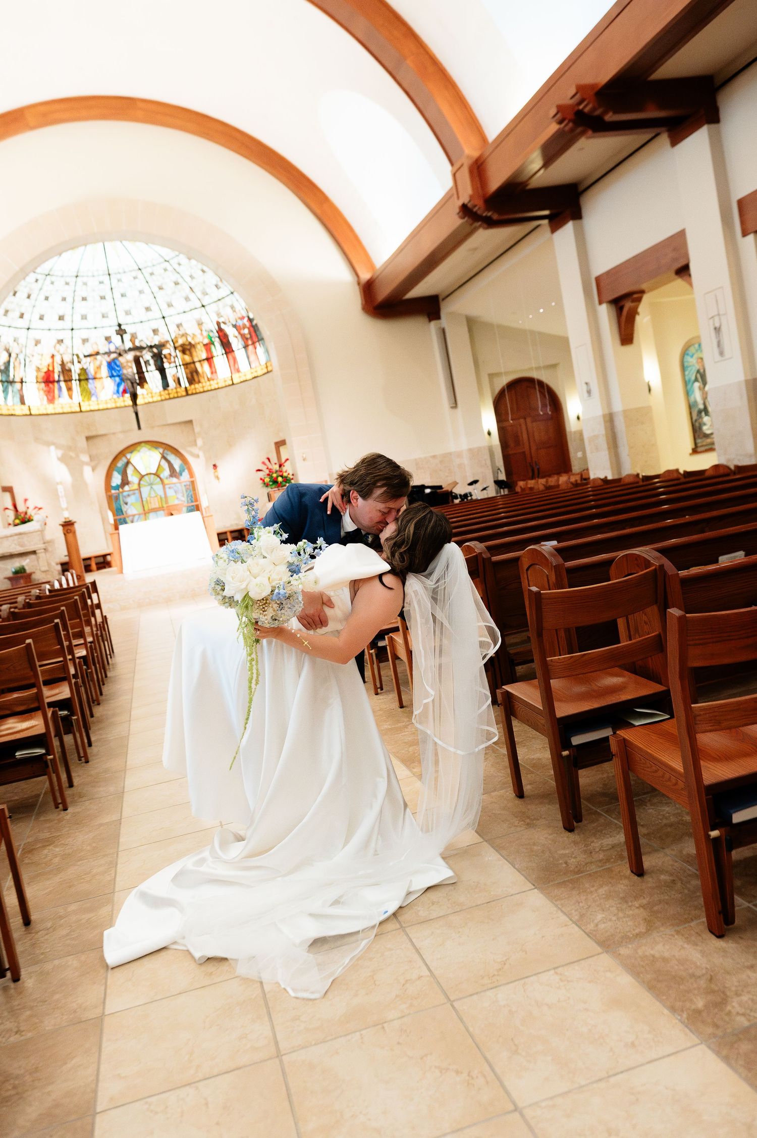 Bride and groom share a romantic kiss in the aisle of a beautiful Catholic church after their wedding ceremony.