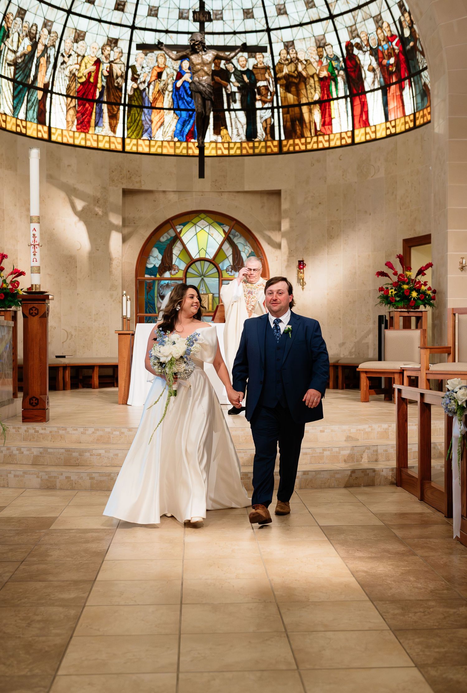 Newlywed couple walks hand-in-hand down church aisle beneath stunning stained glass dome after wedding ceremony.