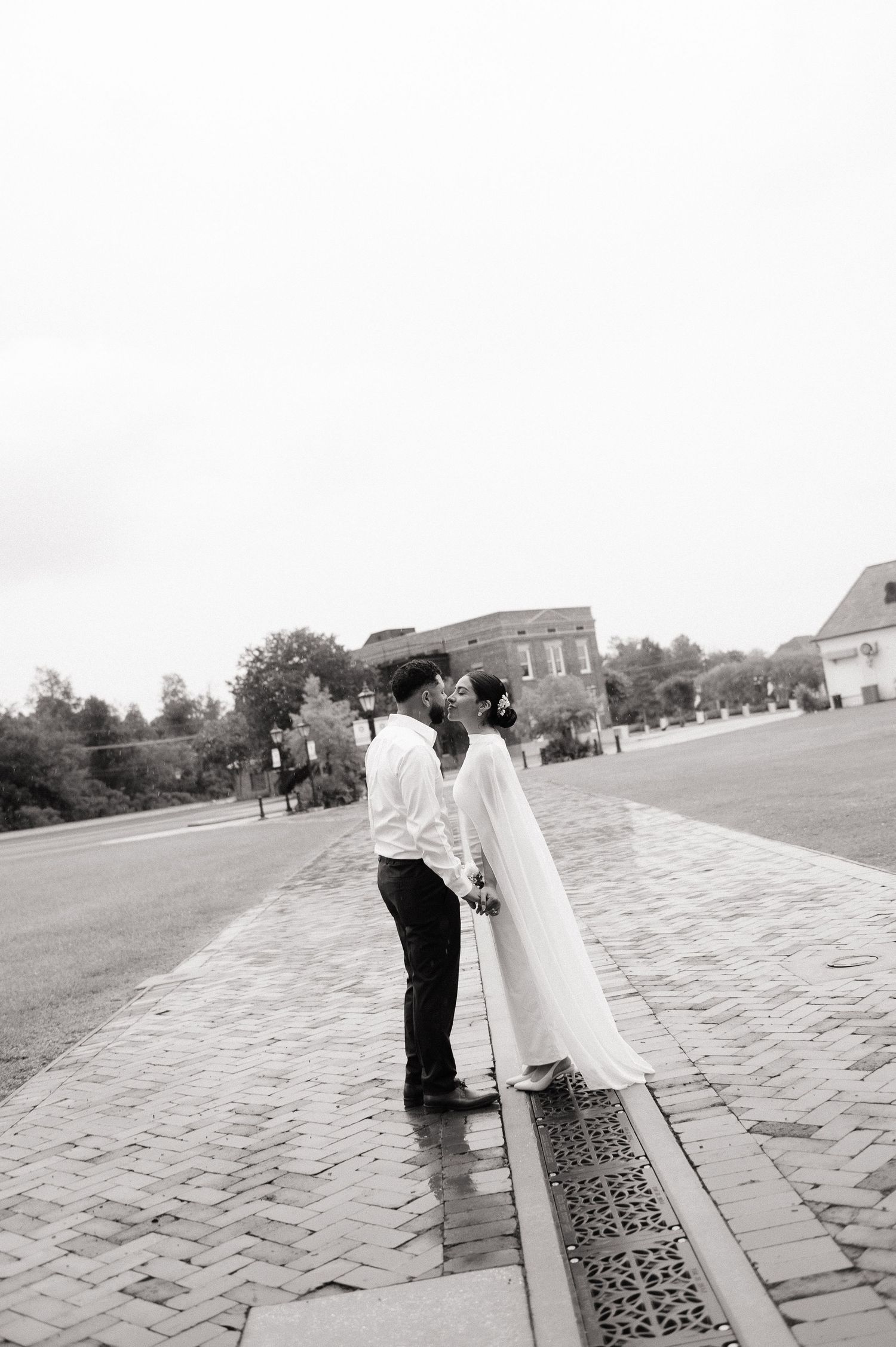 Black and white photo of a couple sharing a kiss in wedding attire on a brick pathway with buildings in the background.