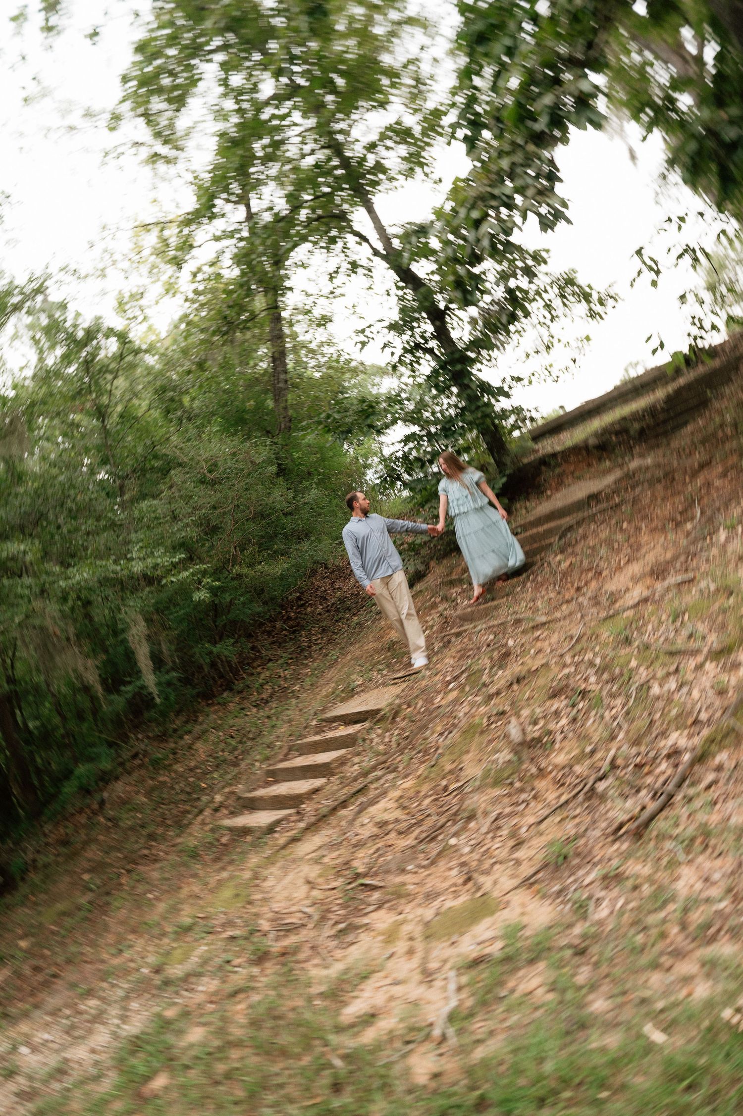 Couple holding hands walking up a wooded hillside path with steps, surrounded by lush green trees.