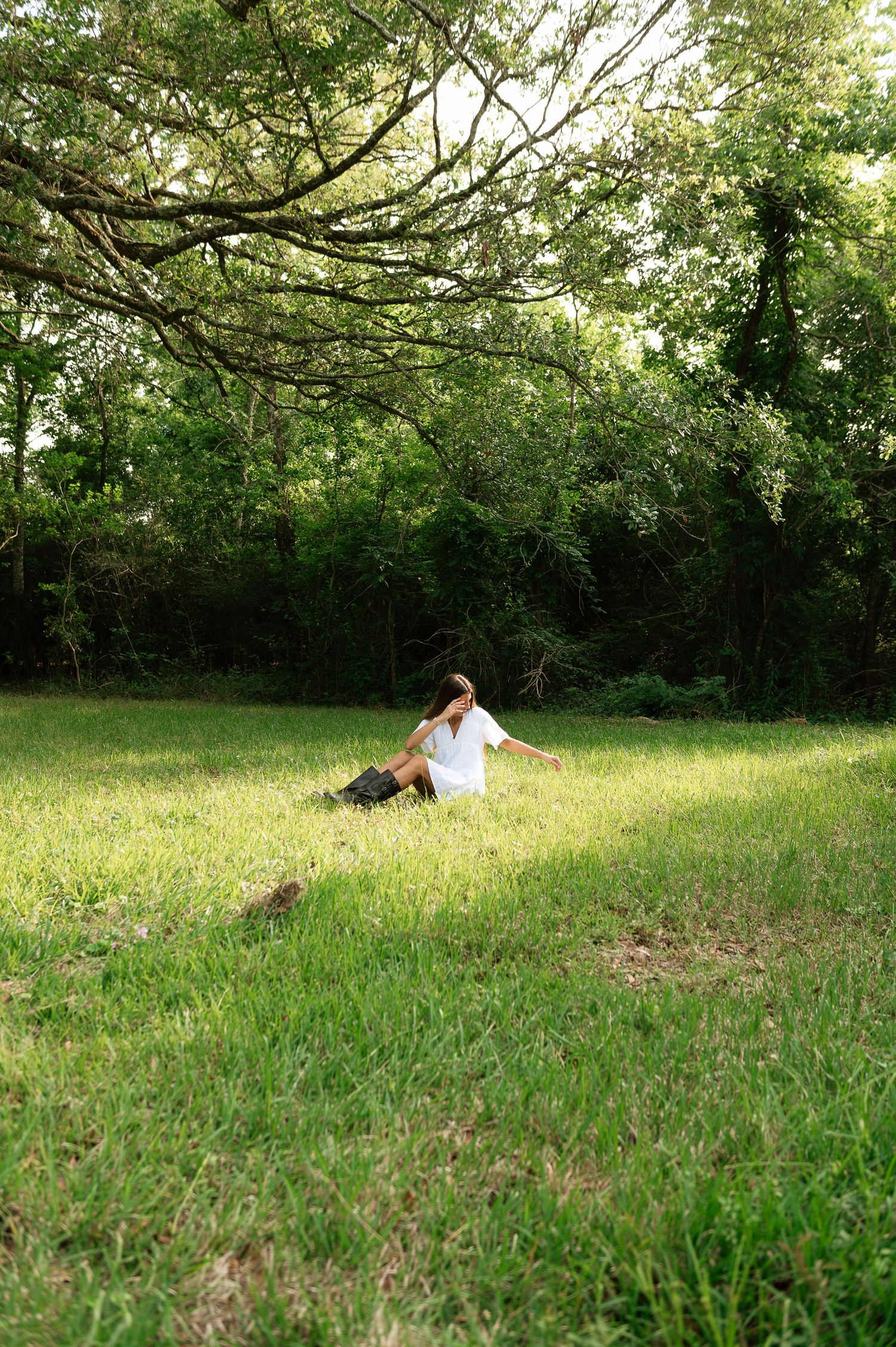A woman in a white top sits on lush green grass in a serene forest clearing with arms outstretched.