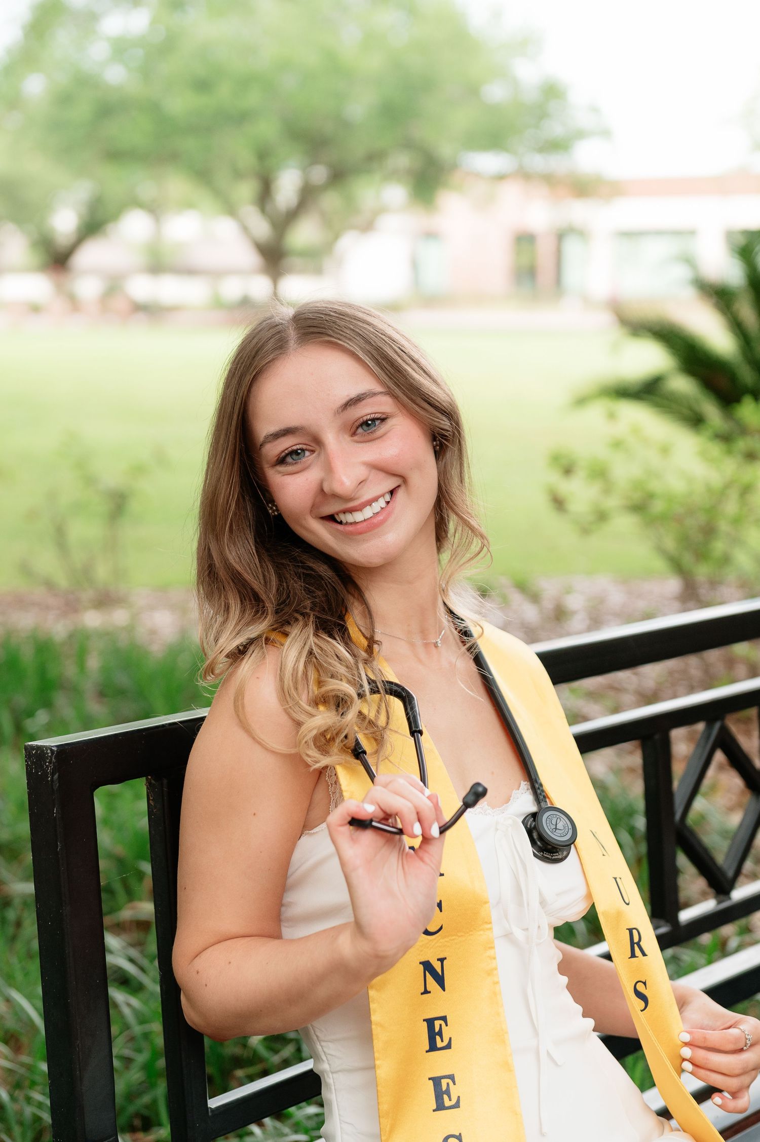 Smiling nursing graduate wearing yellow sash and holding stethoscope at outdoor campus setting.