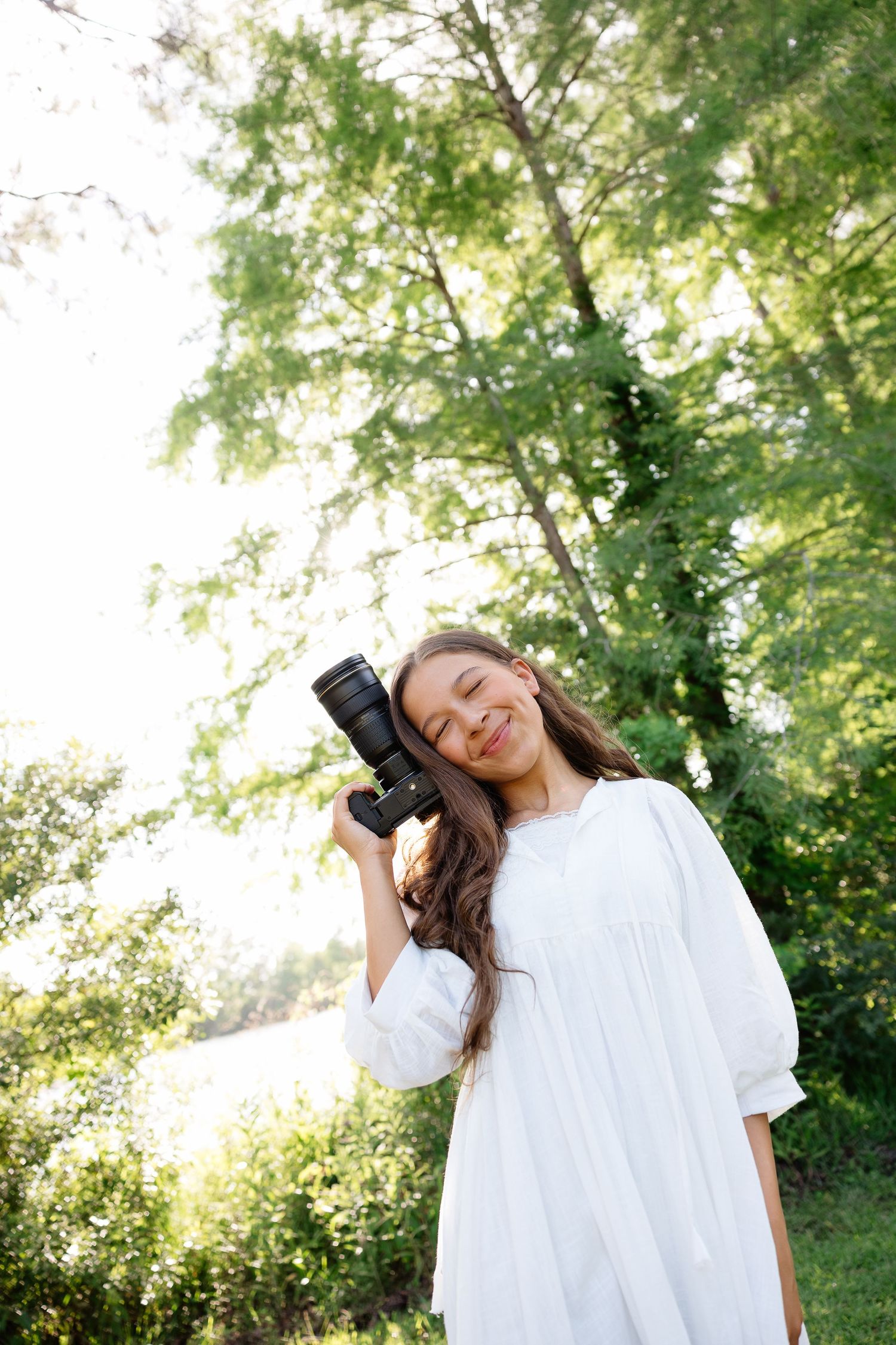 Young girl in white dress smiling while holding a DSLR camera outdoors among green trees in natural light.