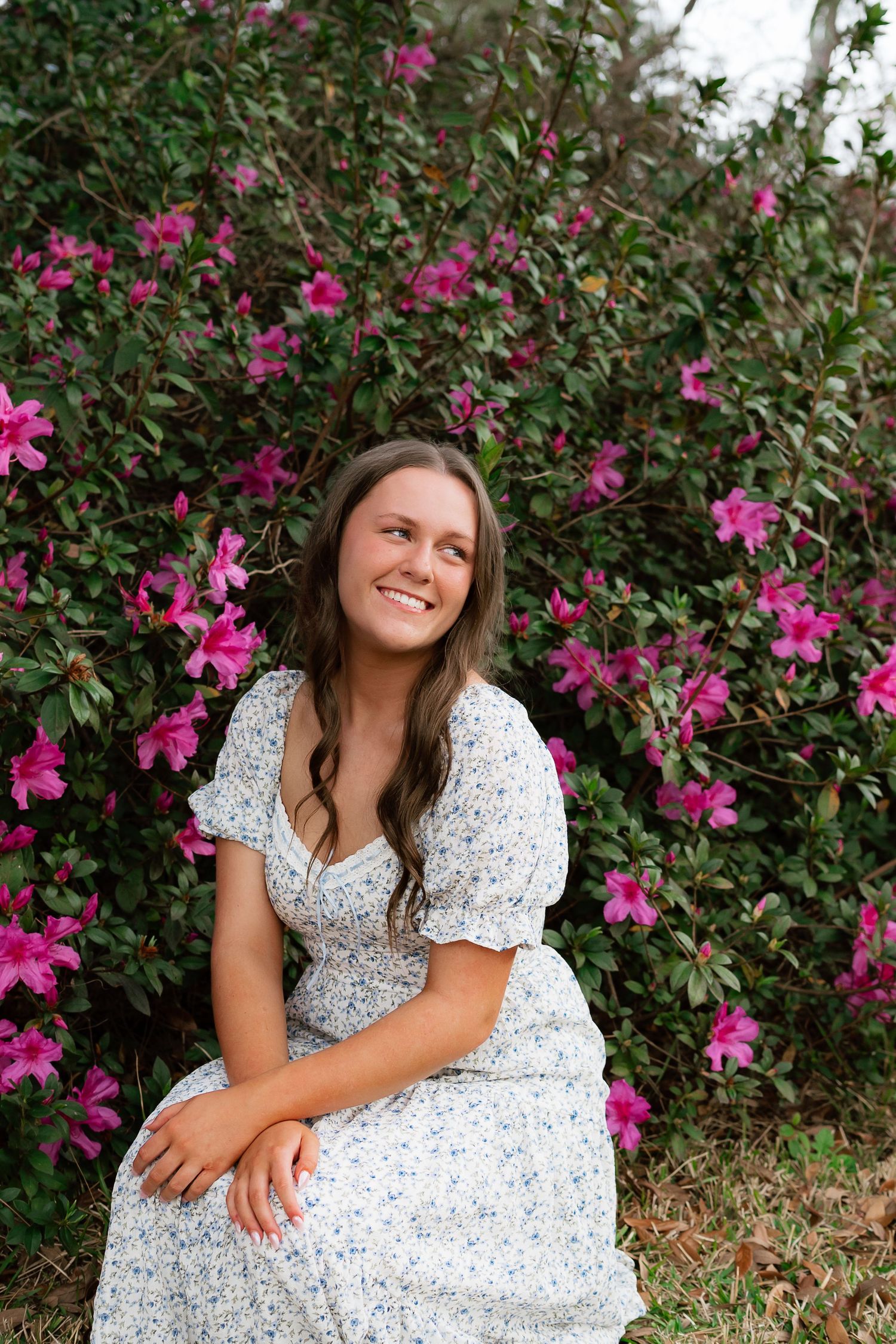 Young woman in floral dress smiling while seated in front of vibrant pink azalea bushes outdoors.